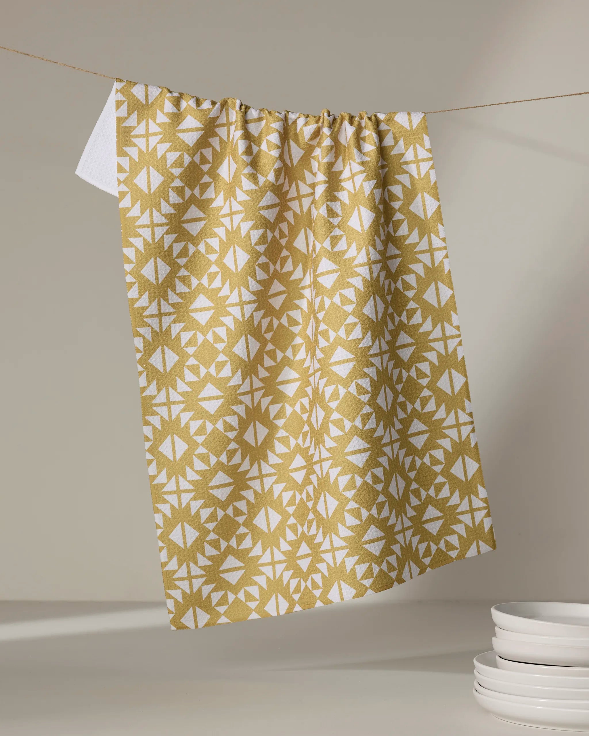 The Ash Cascade "Desert Geo" tea towel, featuring a yellow and white geometric pattern and ultra-absorbent design, hangs on a string near a stack of white plates.