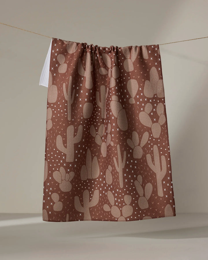 The "Desert Night" tea towel by Rosa Kichline, featuring a beige cactus and star pattern on brown, hangs on a line against a neutral background.