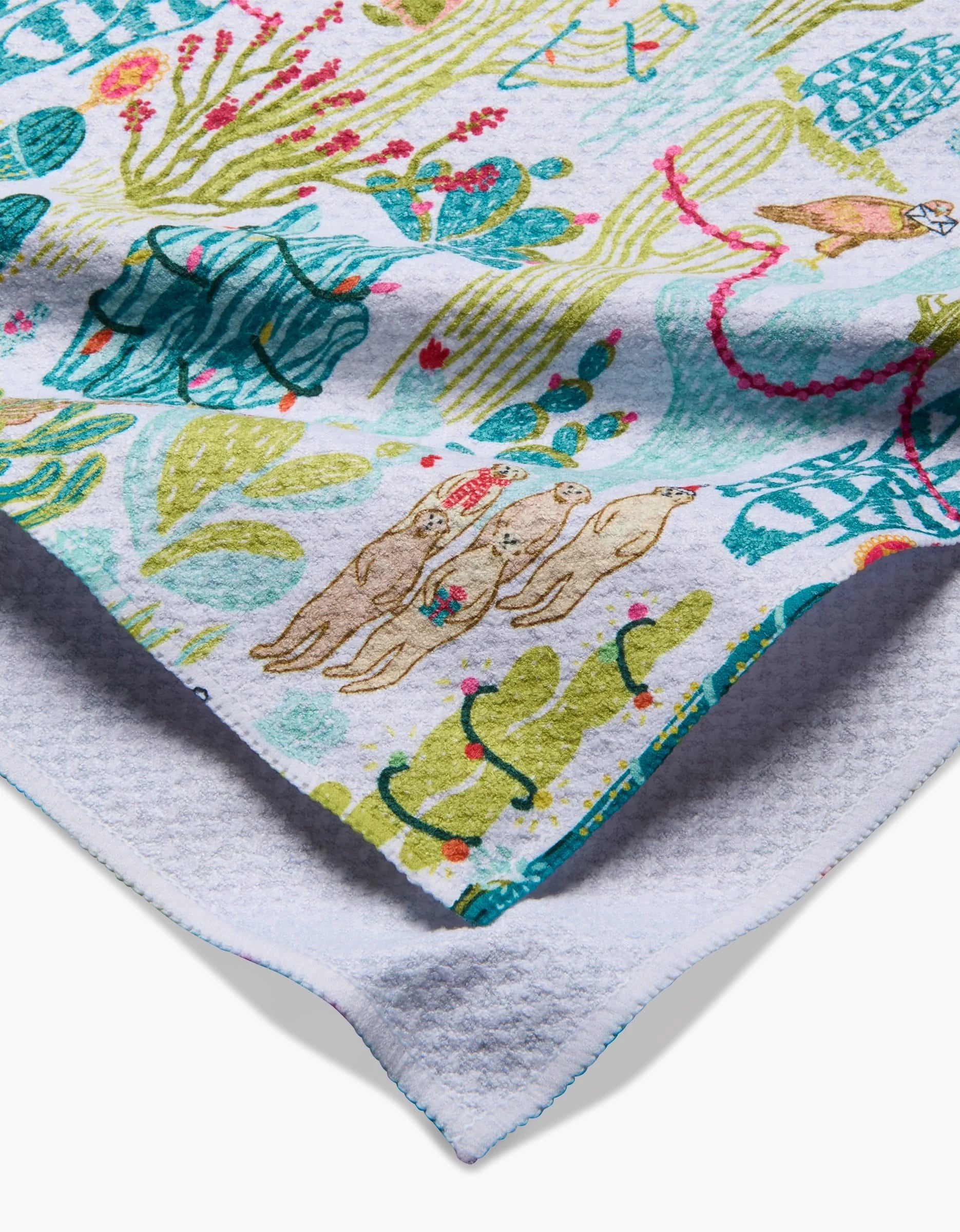 A close-up of Elisabeth Wing’s “Desert On Holiday” eco-friendly kitchen towel, displaying colorful cacti, foliage, and llamas on white. The folded corner reveals its textured, plain underside.