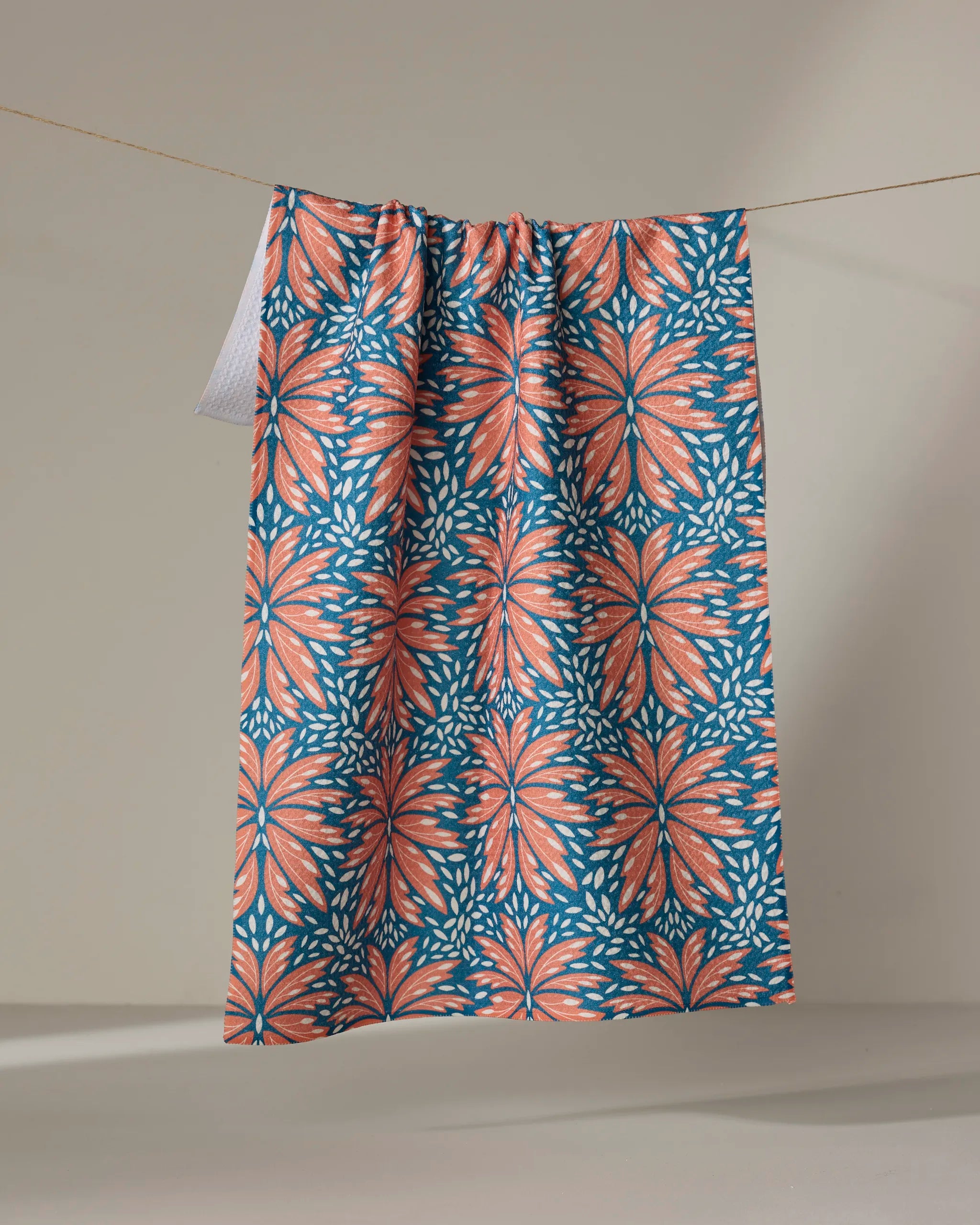 The Dew Drops tea towel by Rebecca Woolbright features a bold floral pattern in coral, blue, and white and hangs over a beige backdrop. Crafted from recycled materials.