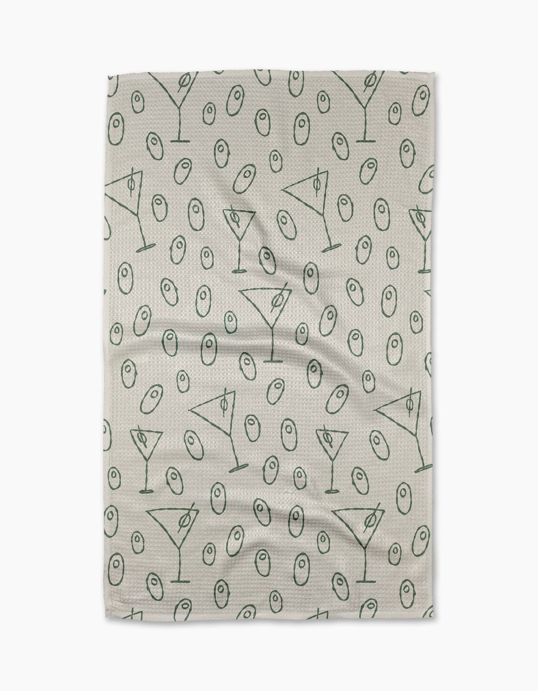 The Dirty Martini tea towel by Rosa Kichline showcases a beige textured background with a repeating pattern of green martini glasses and olives evenly scattered across the surface.