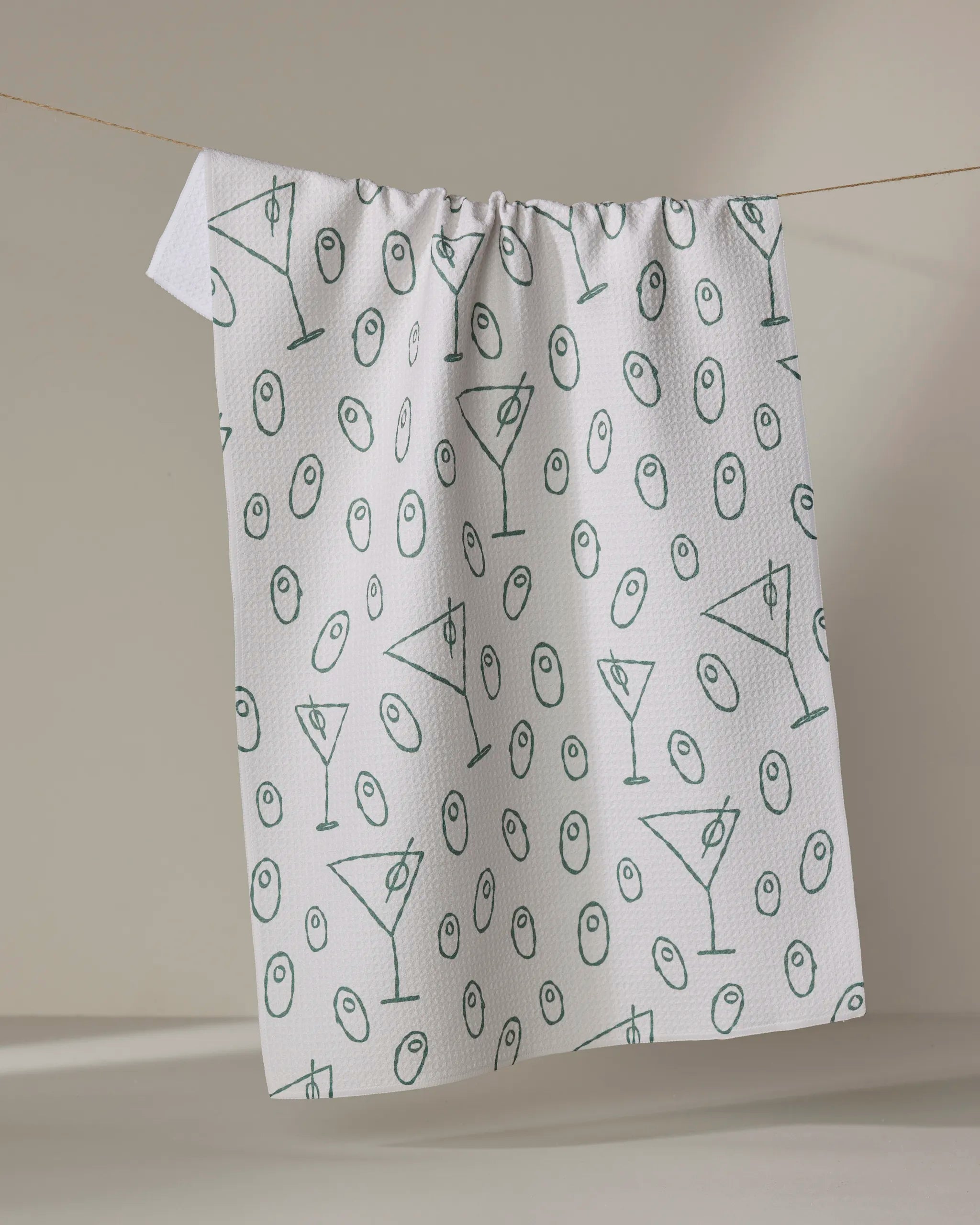 The Rosa Kichline "Dirty Martini" is a white recycled kitchen towel featuring green martini glasses and olives, shown hanging on a clothesline against a neutral background.