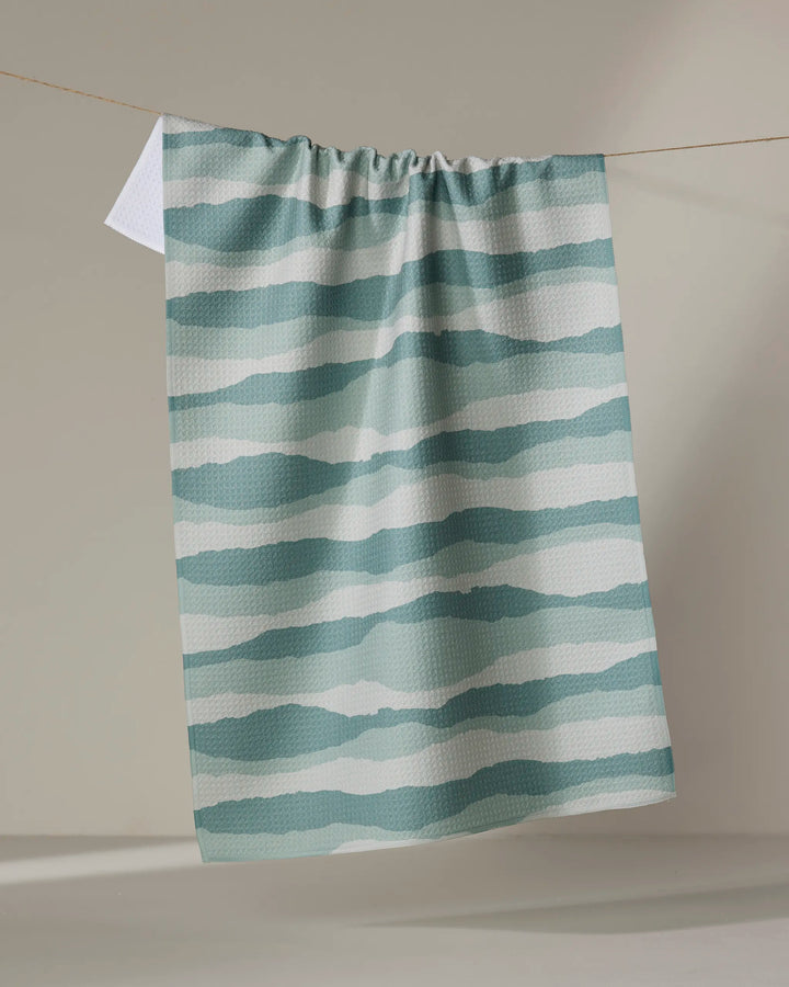 The Distant Landscape tea towel by Twig + Spark, featuring quick-dry fabric and teal and white wavy horizontal stripes, hangs on a clothesline against a neutral backdrop.