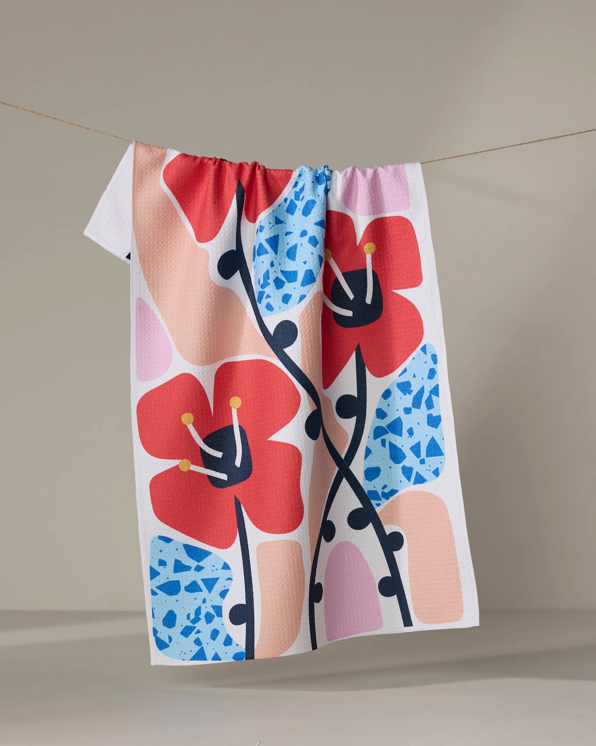 The Dizzy Poppies towel by Happy Hemera, featuring large red flowers, blue accents, and pink-beige blocks in a bold floral design, hangs on a clothesline against a neutral background.