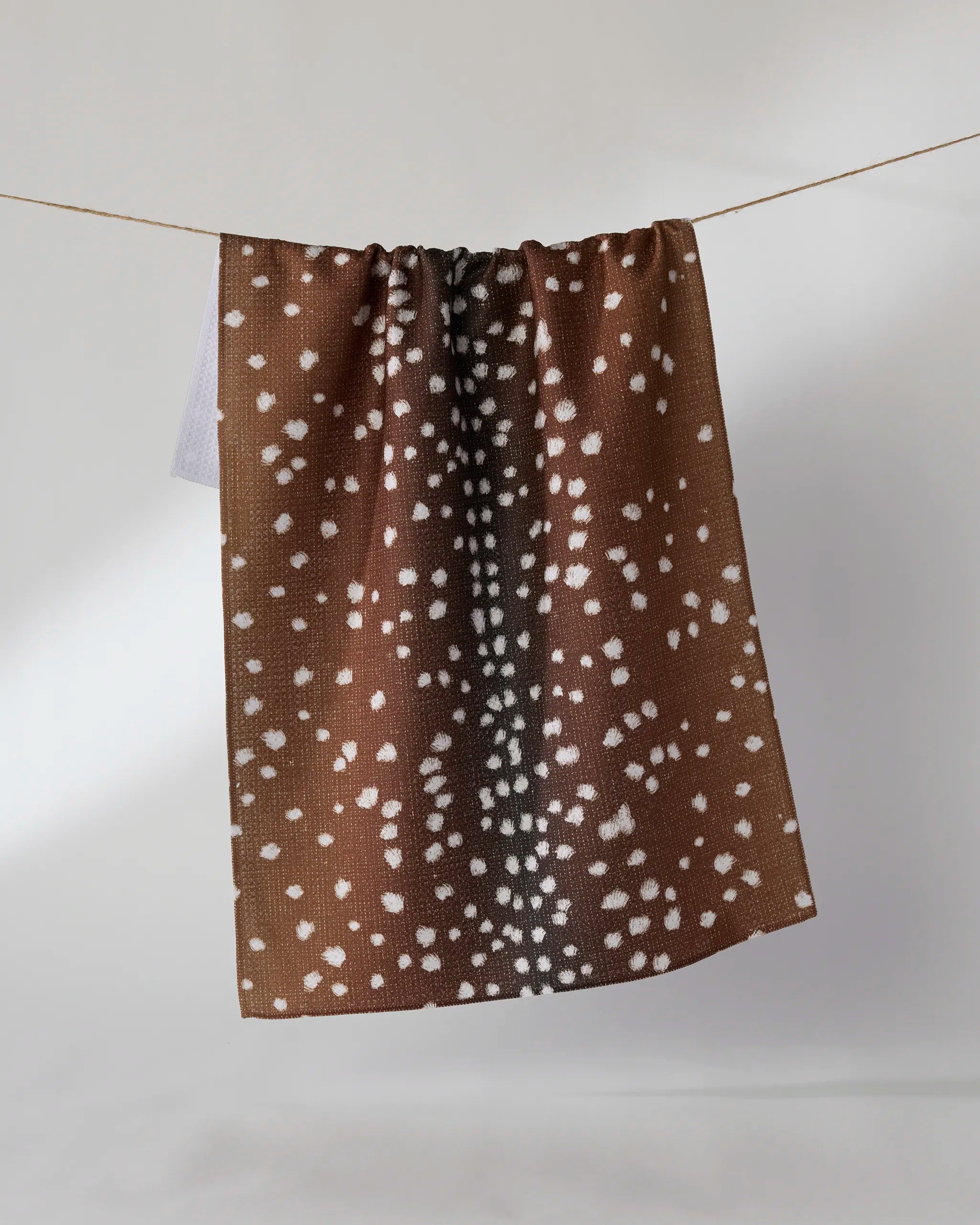 The Doe Trail tea towel by Geometry features white spots on brown, evoking a deer’s coat. Made from recycled materials, this ultra-absorbent towel offers both style and sustainability, shown hanging against a light background.