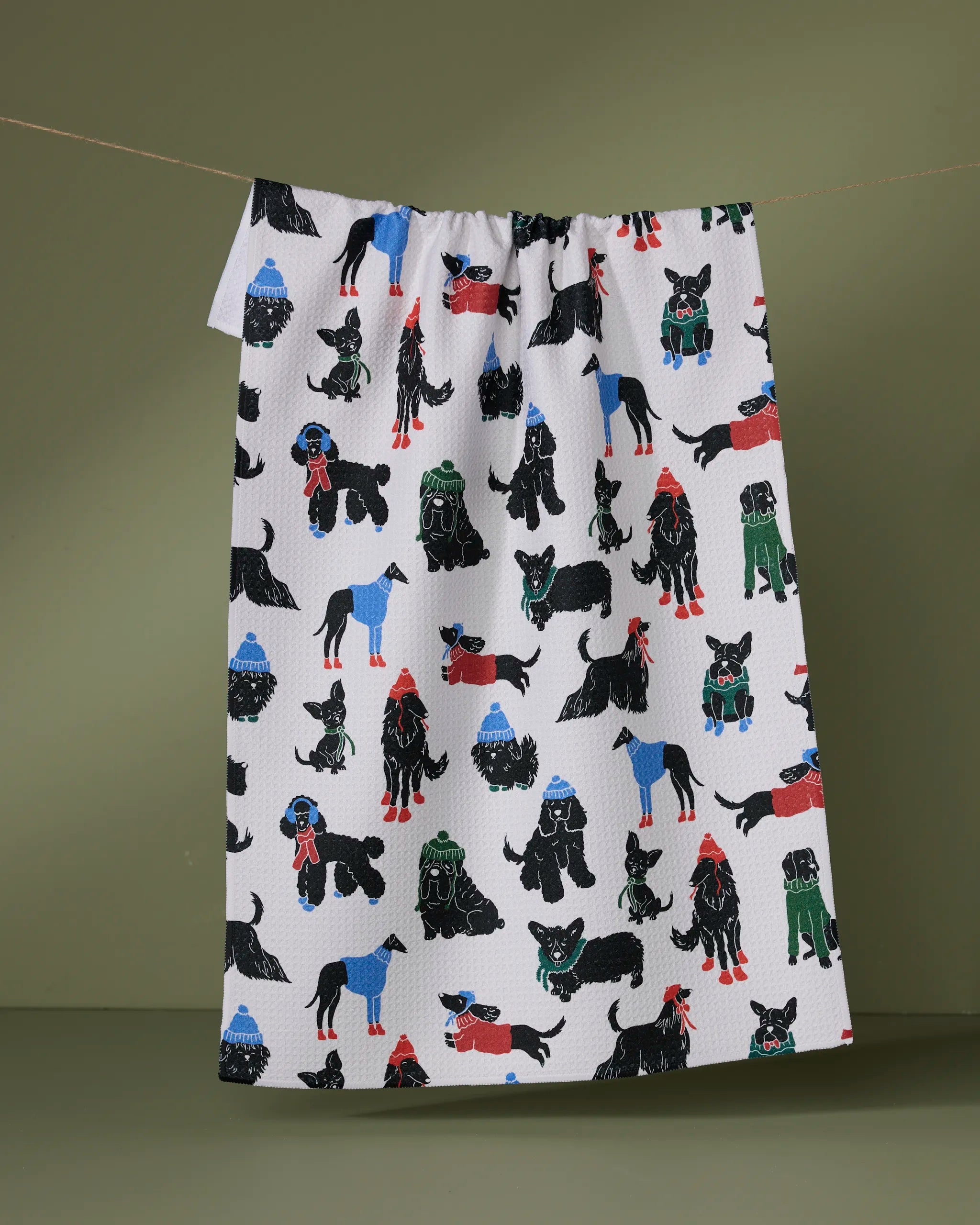 Geometry’s Dogue Magazine tea towel is ultra-absorbent, made from recycled materials, and showcases vibrant illustrations of dogs in black, red, blue, and green hanging on a string against an olive-green background.