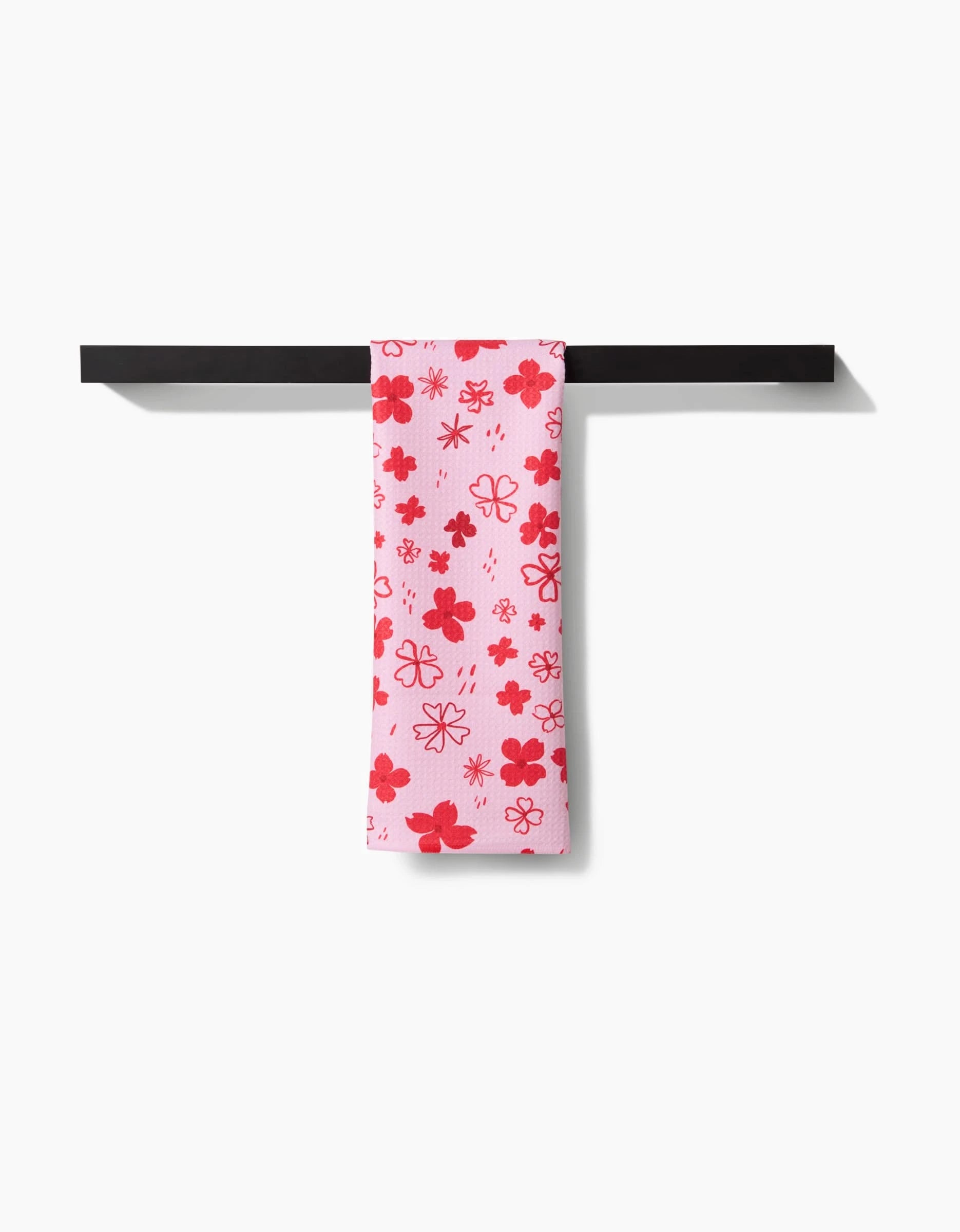 The Dogwood Hearts tea towel by Elana Gabrielle, featuring red and pink floral patterns on white and made from recycled materials, hangs neatly over a black towel bar against a white background.