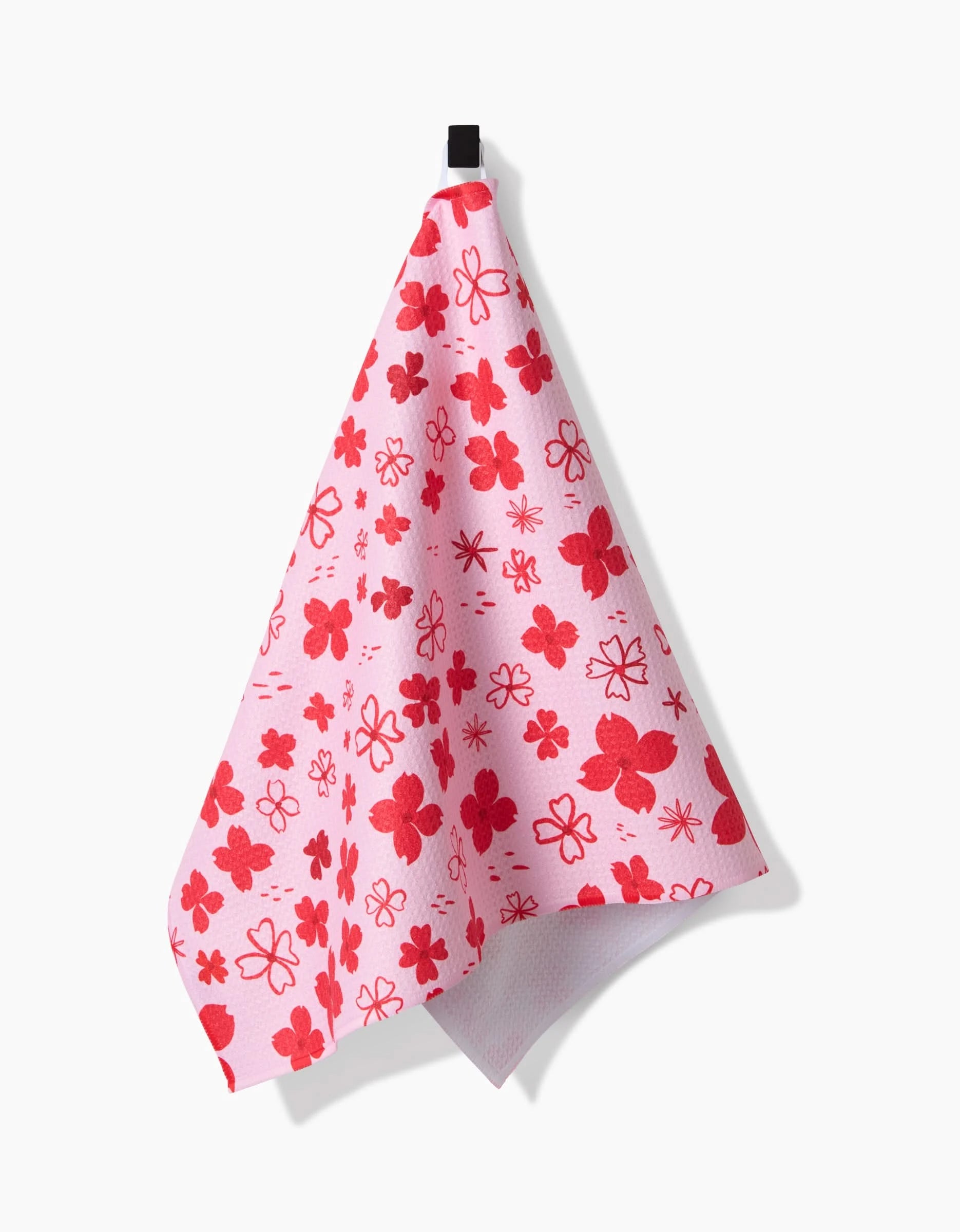 The Dogwood Hearts tea towel by Elana Gabrielle, featuring red and pink flower patterns, hangs from a black hook against a white background.