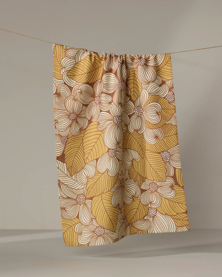 The Dogwood kitchen towel by Katie O'Shea features bold mustard yellow, brown, and white floral patterns with large overlapping flowers, hanging from a string against a neutral background.