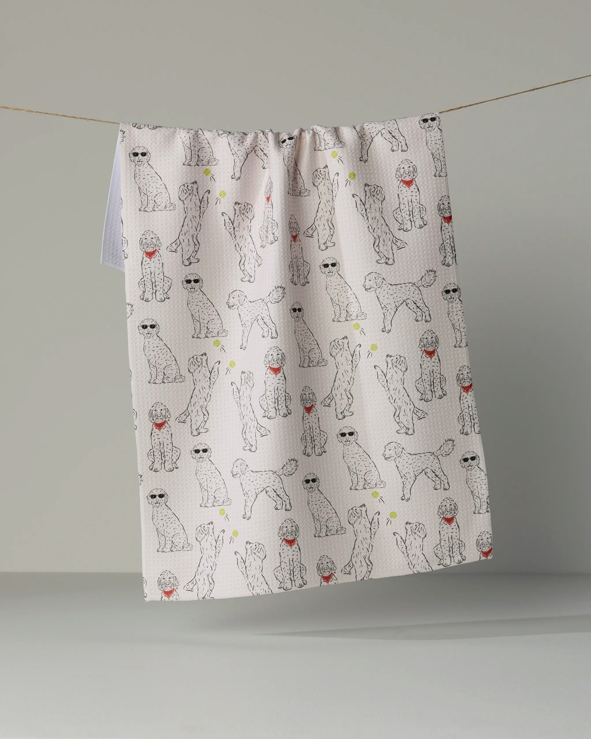 A light-hued Doodle Daze tea towel by Geometry hangs on a line, showcasing a whimsical design of cartoon dogs sporting sunglasses and red hats atop a subtle polka-dot backdrop. Made from recycled materials, this delightful kitchen towel brings eco-friendly flair and playful charm to any space.