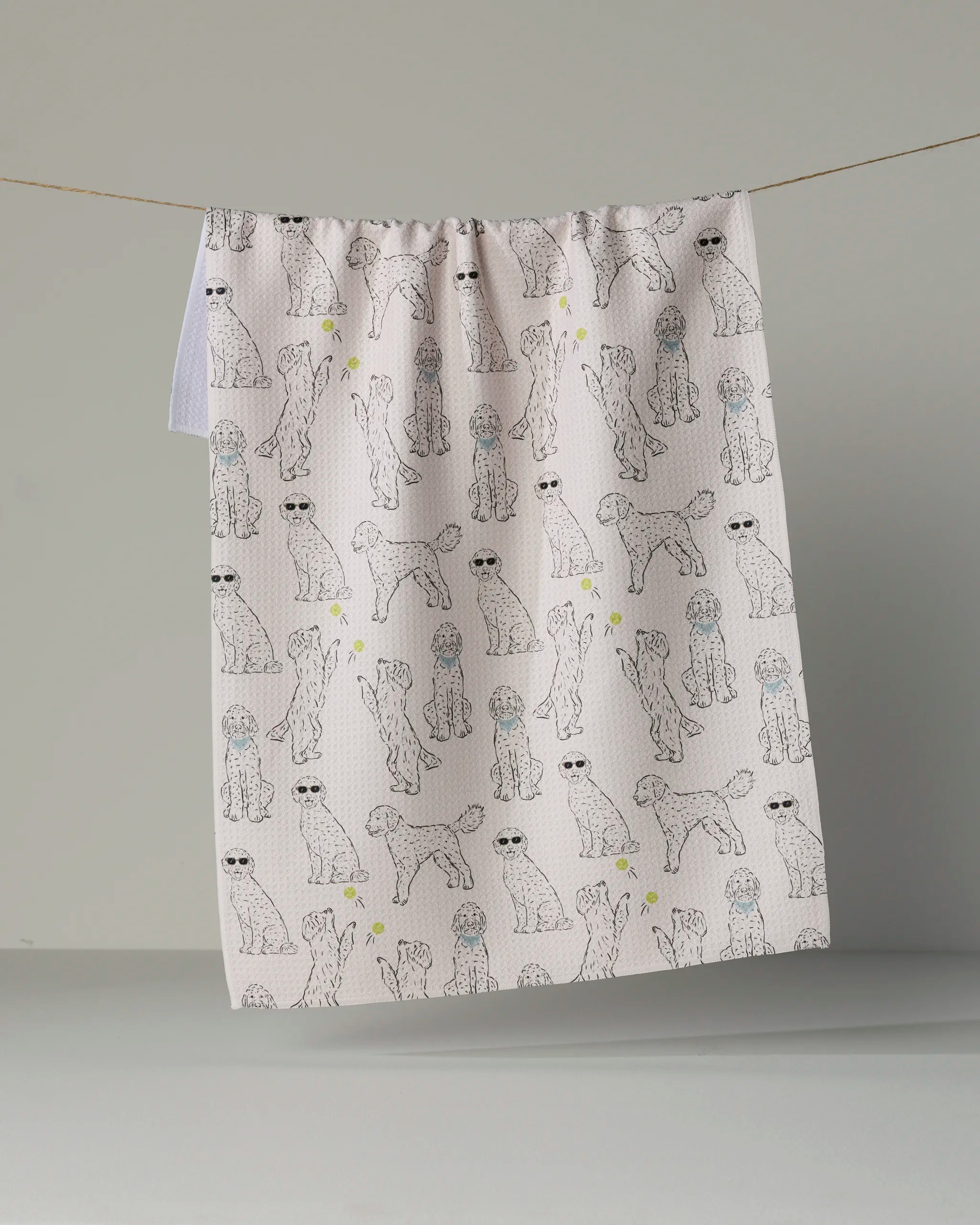 The Geometry “Doodle Daze” tea towel, featuring a white background with a repeating pattern of illustrated poodles—some sporting sunglasses—hangs on a line against a plain gray backdrop.