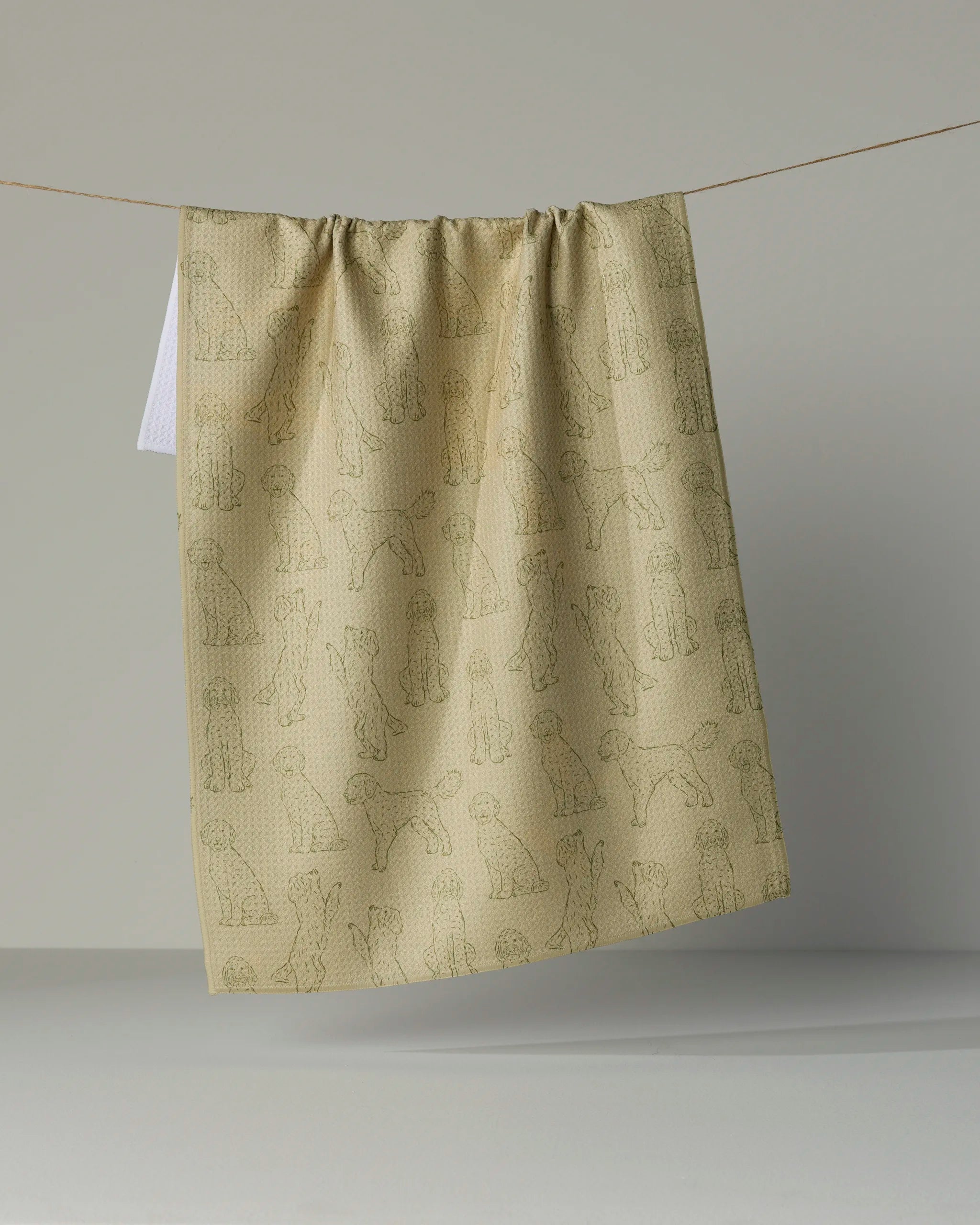 A “Doodle Pups” recycled kitchen towel by Geometry, featuring an outline pattern of various dogs in beige, hangs on a clothesline against a plain, light gray background.