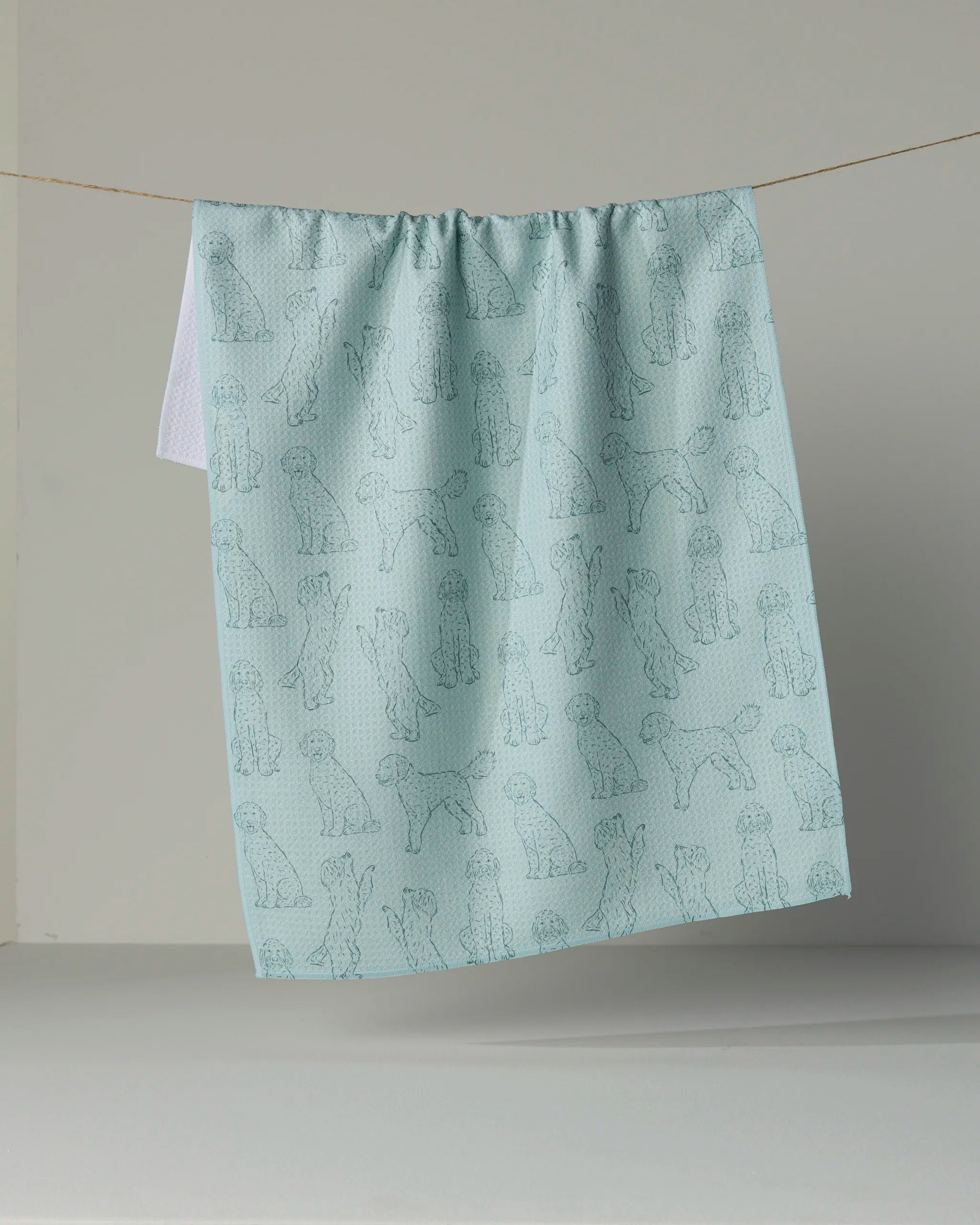 A light blue “Doodle Pups” recycled kitchen towel by Geometry, featuring outlines of various dog breeds, hangs on a string against a plain background.