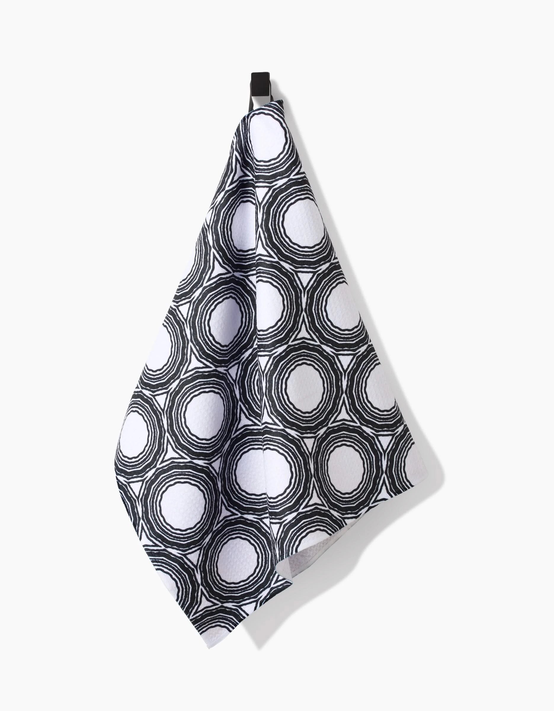 The Katherine Berkeley Dosido Tea Towel, made from Geoweave fabric for superior absorbency, features a black and white concentric ring pattern and hangs from a black hook against a plain white background.