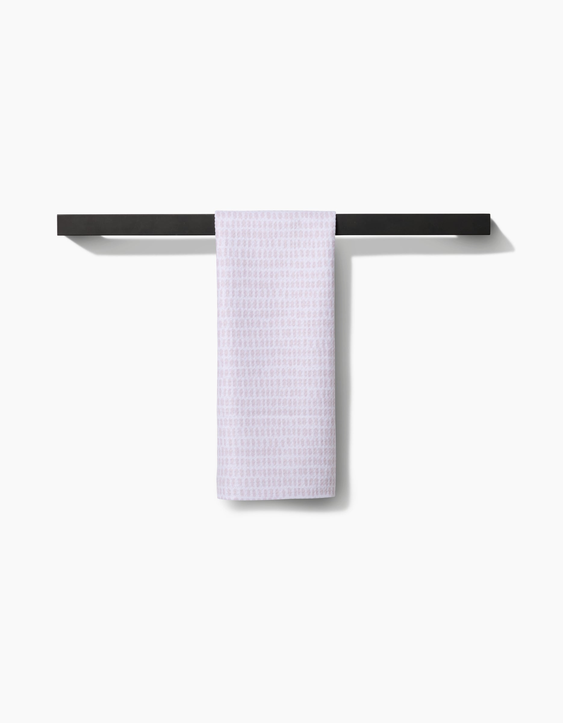 The Geometry Dot Dot Dot Blushed Bergamot towel, ultra-absorbent and white, is neatly draped over a minimalist black towel rack on a white wall, casting a subtle shadow to the right.