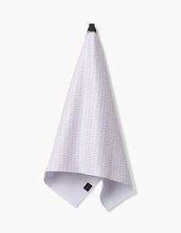 The Dot Dot Dot Blushed Bergamot tea towel by Geometry, made from recycled materials, hangs from a black hook on a white background. It showcases a light gray textured weave with a subtle pattern and has a black label near the bottom.