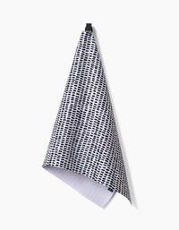 The Geometry Dot Dot Dot Fresh Linen ultra-absorbent tea towel, featuring a black and white dotted grid pattern, hangs from a black hook against a plain white background.