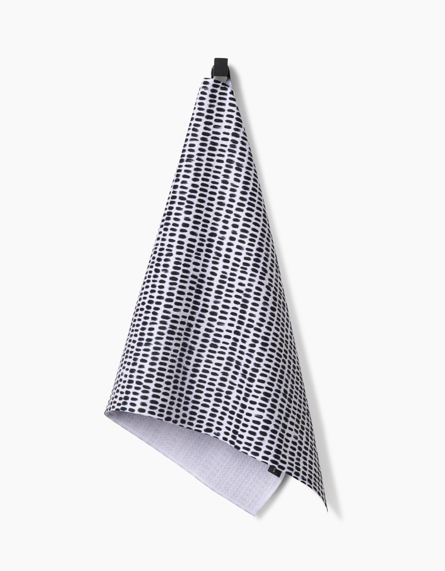 The Geometry Dot Dot Dot Fresh Linen ultra-absorbent tea towel, featuring a black and white dotted grid pattern, hangs from a black hook against a plain white background.