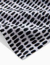 A close-up of Geometry’s Dot Dot Dot Fresh Linen tea towel shows its black abstract oval dot pattern on white, emphasizing the ultra-absorbent fabric’s texture and folds.