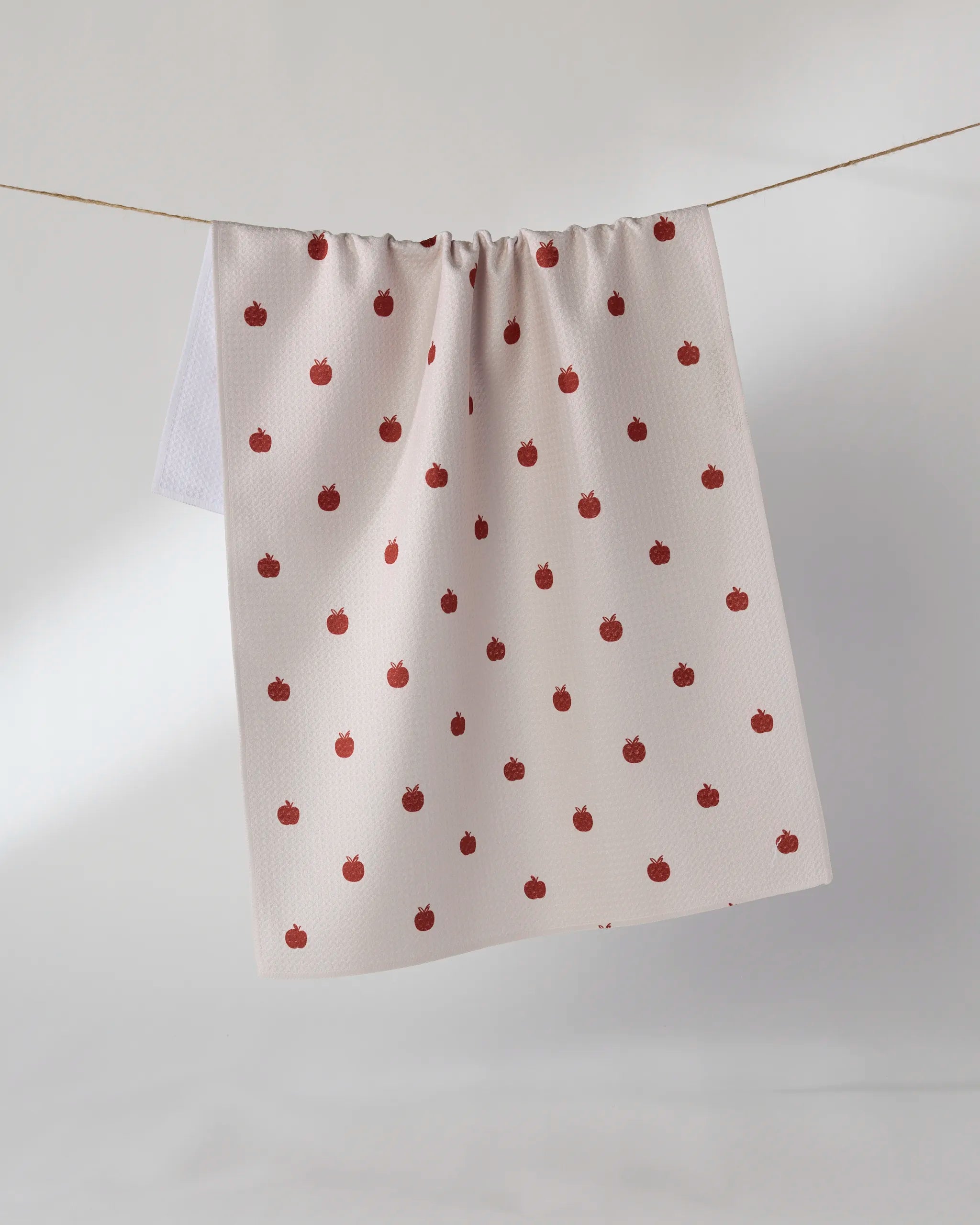 The Geometry Dotty Apple kitchen tea towel, crafted from recycled materials and adorned with a playful red apple pattern, hangs on a clothesline against a neutral background.