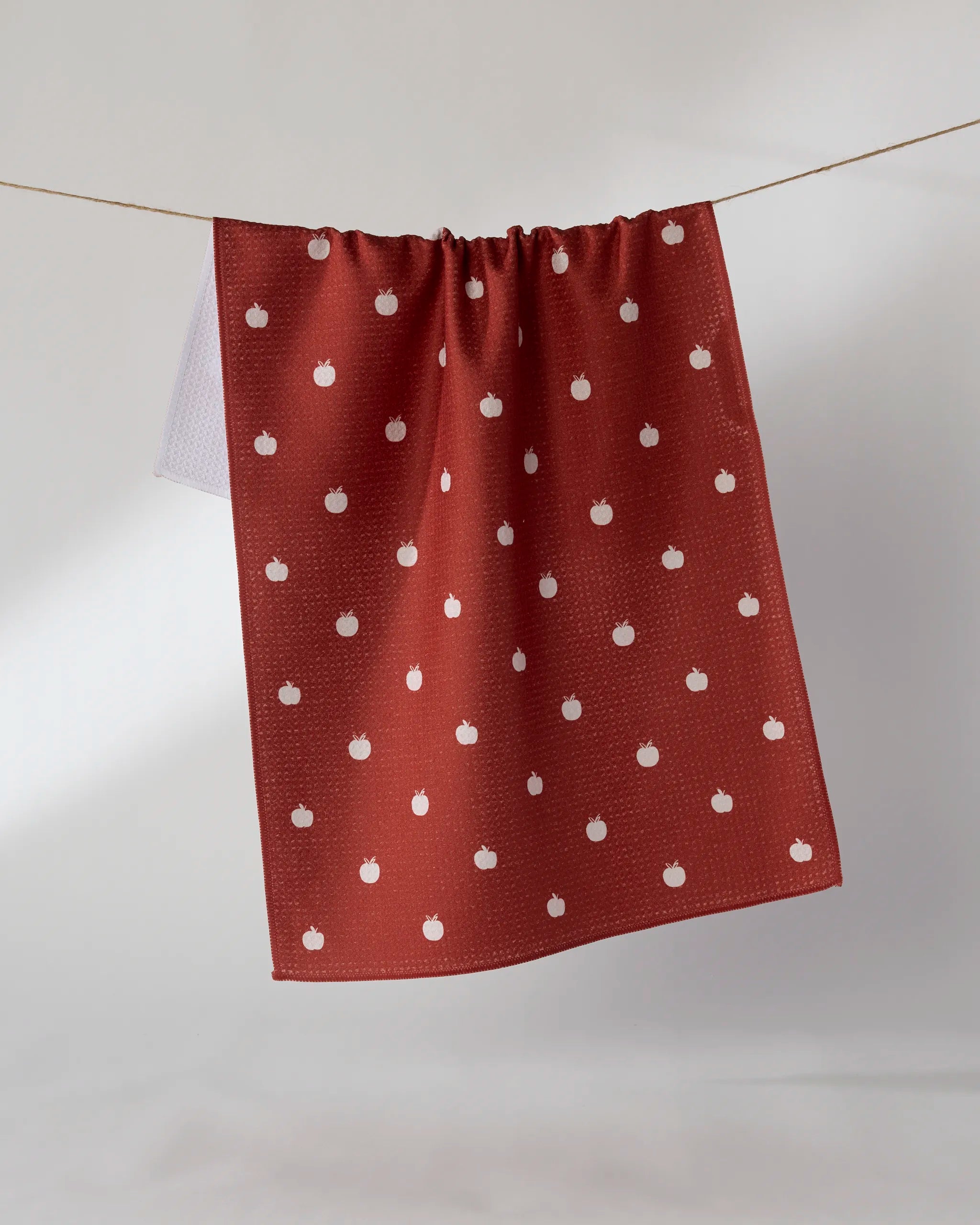 The Geometry Dotty Apple tea towel, featuring a small white apple pattern on red and made from recycled materials, hangs on a clothesline against a plain, light background.