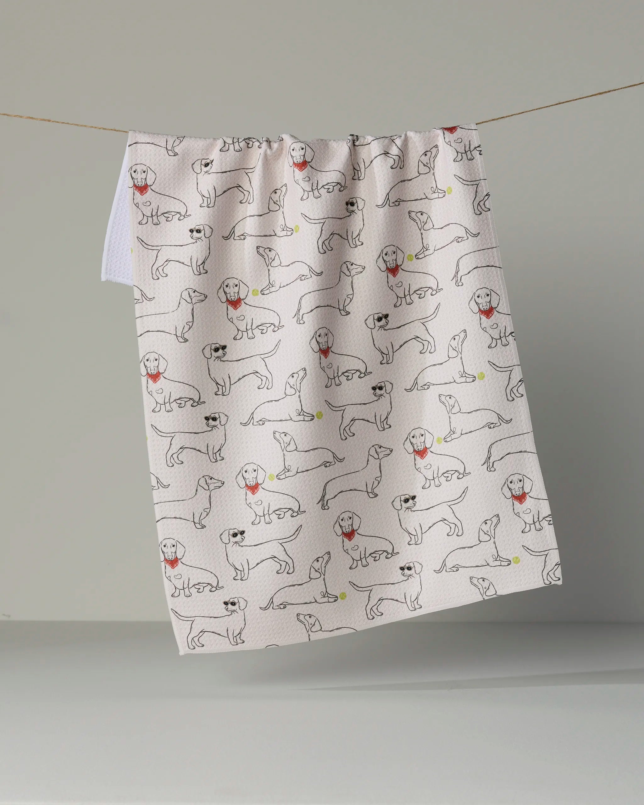 The “Doxie Pals” tea towel by Geometry, featuring a lively pattern of outlined dogs in different poses—some with red collars and some chasing yellow tennis balls—hangs on a clothesline against a simple backdrop. This white, eco-friendly kitchen towel is sure to add charm and personality to any space.
