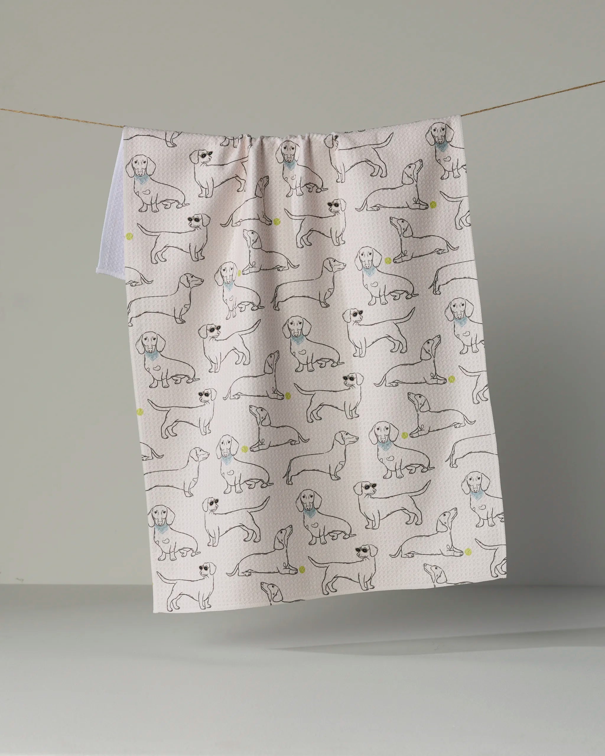 The Doxie Pals tea towel by Geometry, crafted from eco-friendly materials, showcases a delightful pattern of hand-drawn dachshunds—some with playful yellow balls—displayed against a plain gray background.