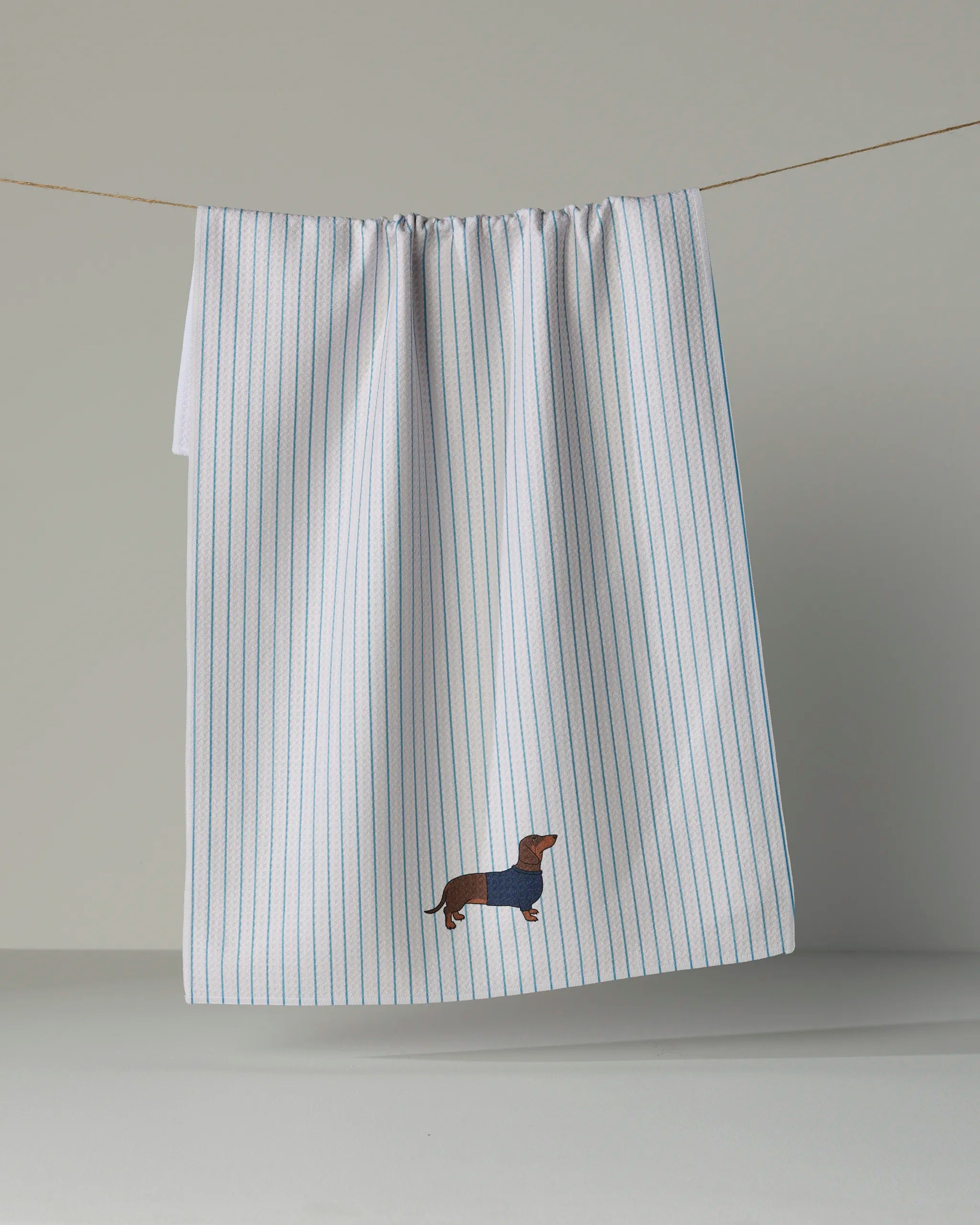 A white Geometry Doxie Stripe kitchen tea towel with thin blue stripes hangs on a line. Ultra-absorbent, it features an embroidered brown dachshund in a blue sweater near the bottom edge.
