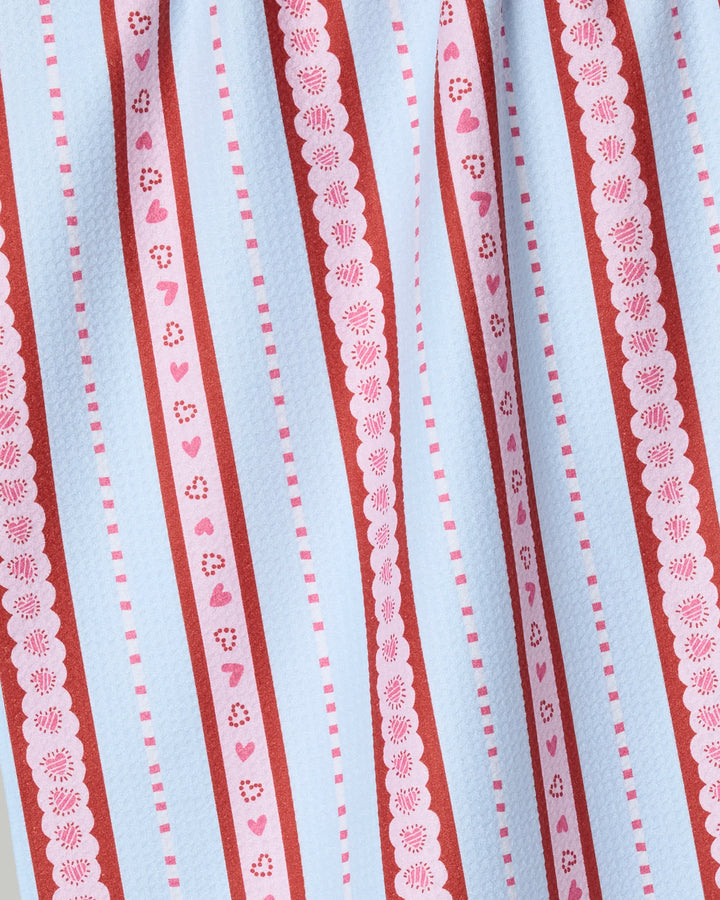 The Dreaming of You tea towel by Geometry features white Geoweave fabric with vertical red and pink stripes, hearts, flowers, scalloped edges, and geometric designs. Softly pleated, it's made from recycled materials.