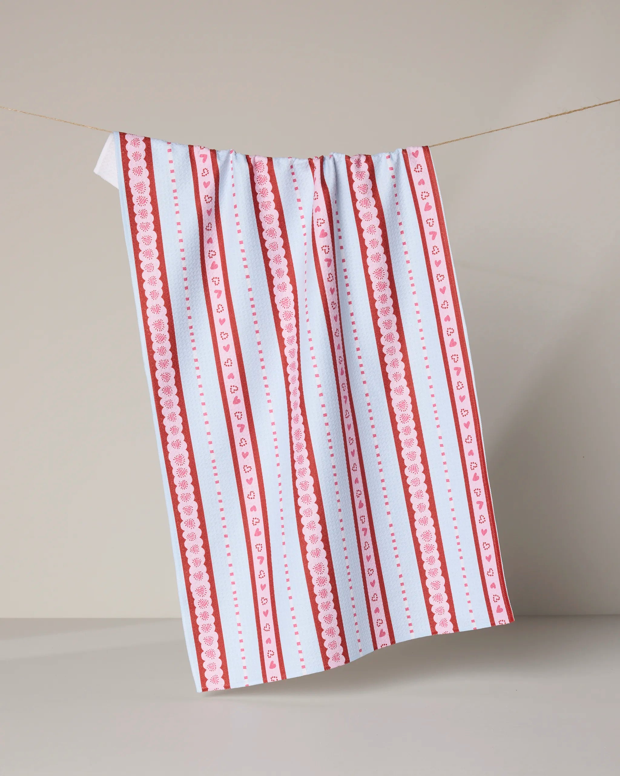 The Geometry "Dreaming of You" tea towel, with quick-drying fabric and vertical red, pink, and white stripes adorned with heart and floral designs, hangs on a clothesline against a neutral background.