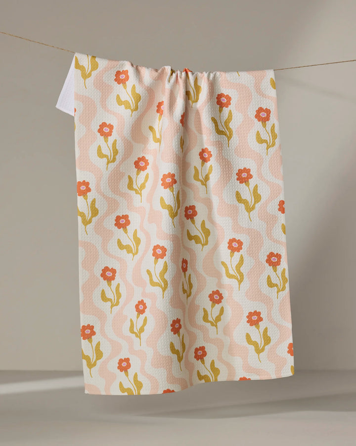 The Dreamy Path tea towel by Gerdadzy, featuring an abstract floral pattern in orange, yellow, and pink, hangs from a string against a neutral background.