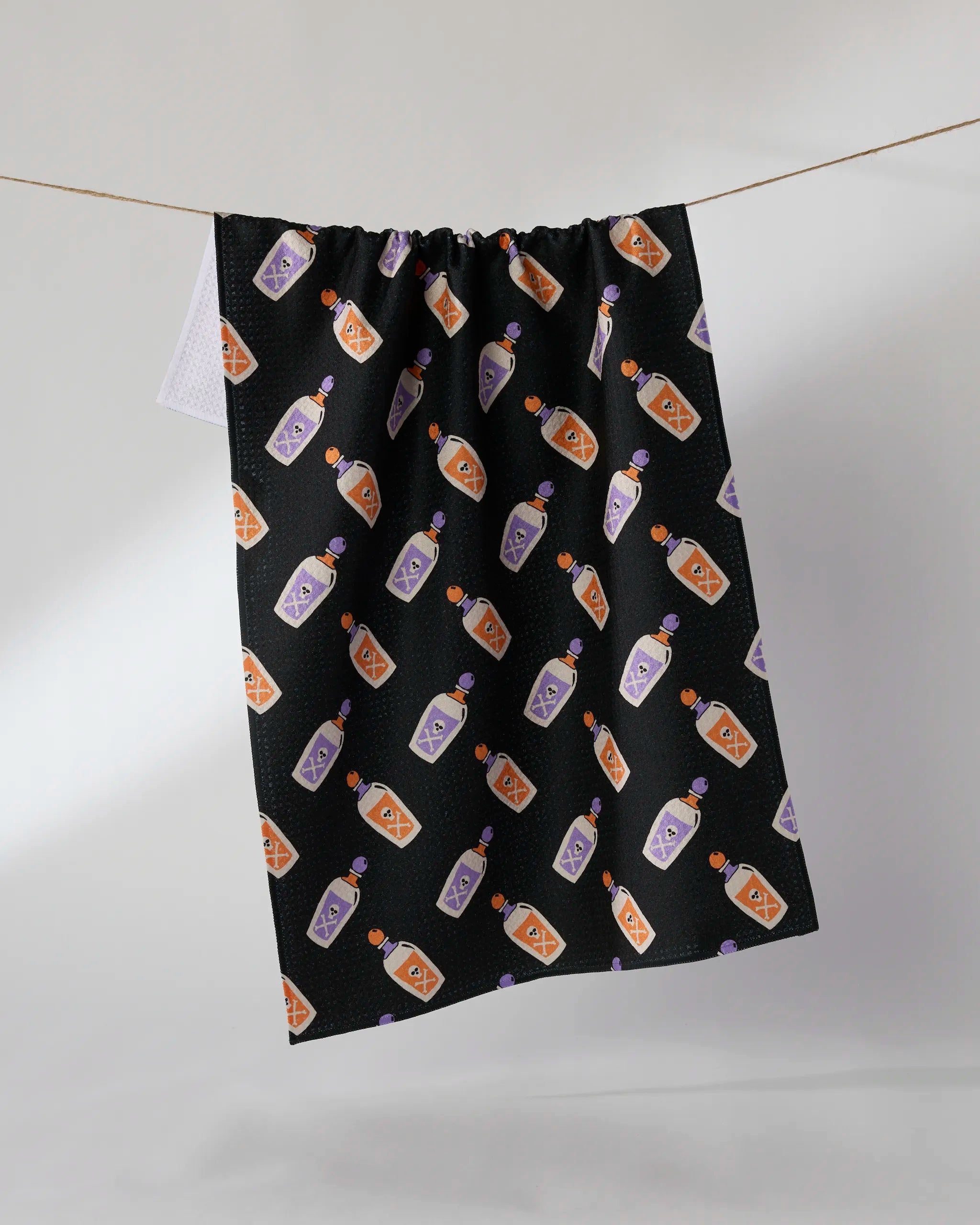 The Geometry "Drink Me" tea towel is ultra-absorbent and crafted from recycled materials, featuring a repeating pattern of whimsical potion bottles in orange and purple on black, shown hanging against a plain backdrop.
