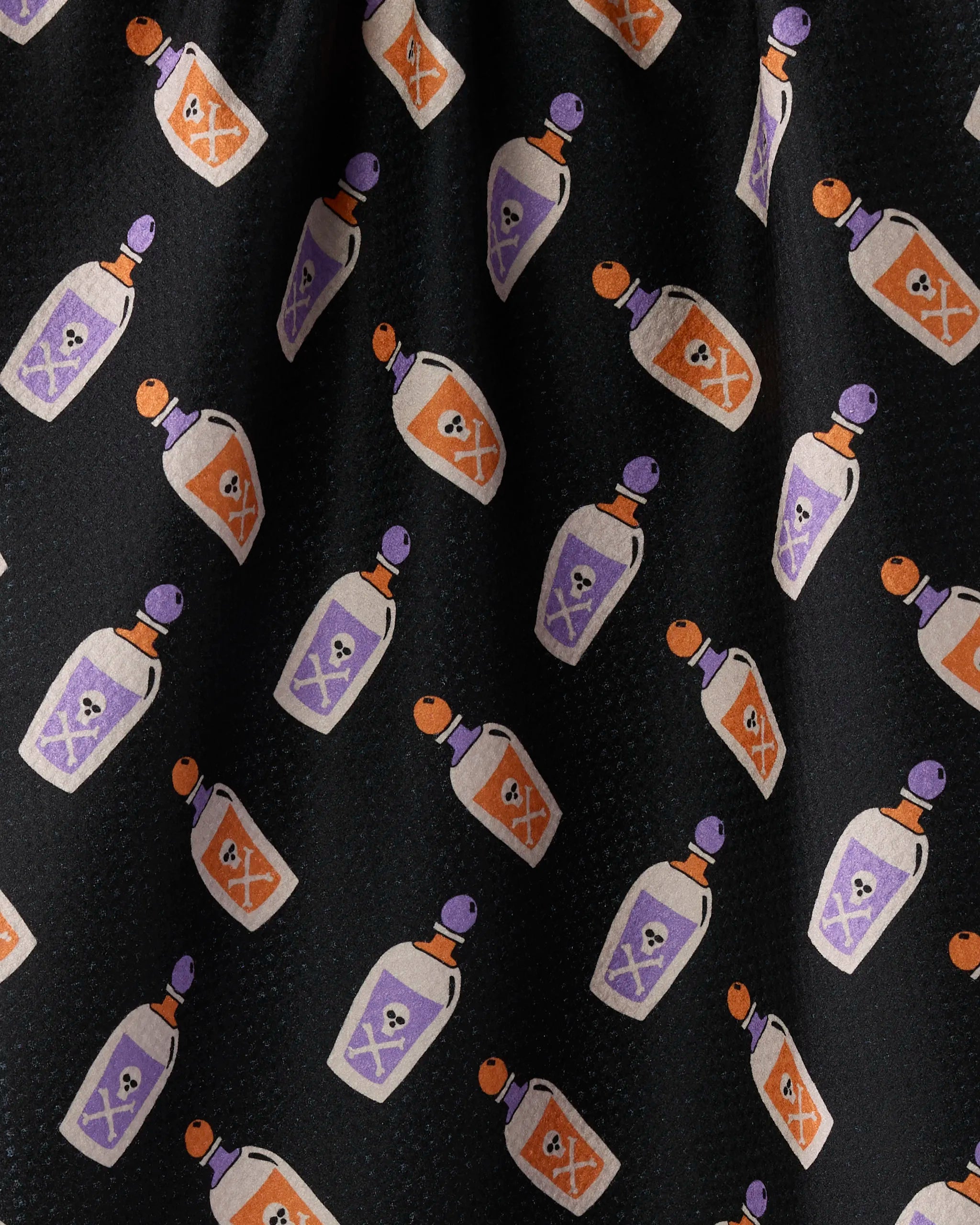 The "Drink Me" tea towel by Geometry features small orange and purple potion bottles with white skulls on black, creating a whimsical, spooky vibe. Made from ultra-absorbent recycled materials.