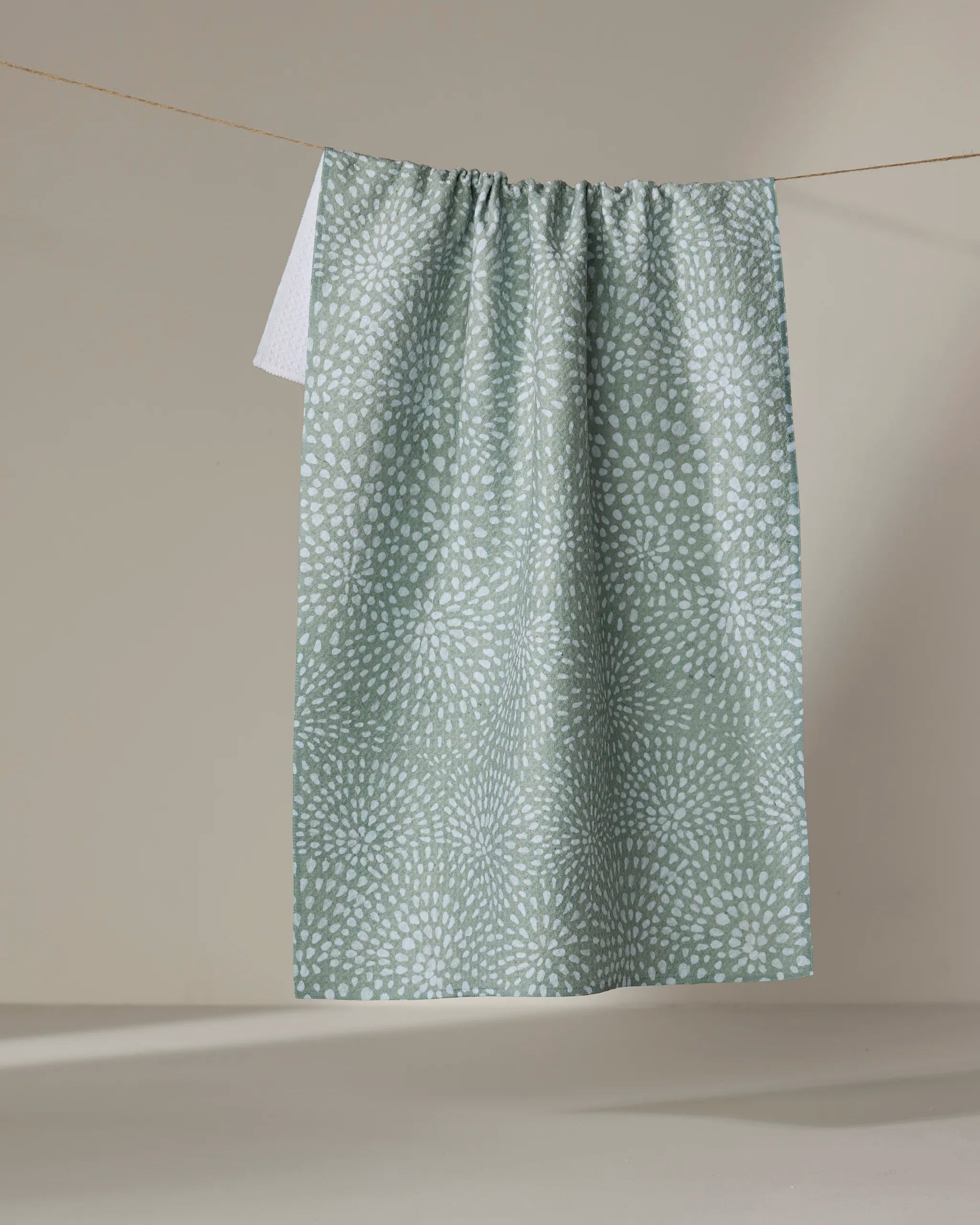 The Droplets tea towel by Katie O'Shea, featuring ultra-absorbent light green fabric with a white dotted pattern, hangs on a clothesline against a plain beige background. A corner of a white towel peeks out behind it.