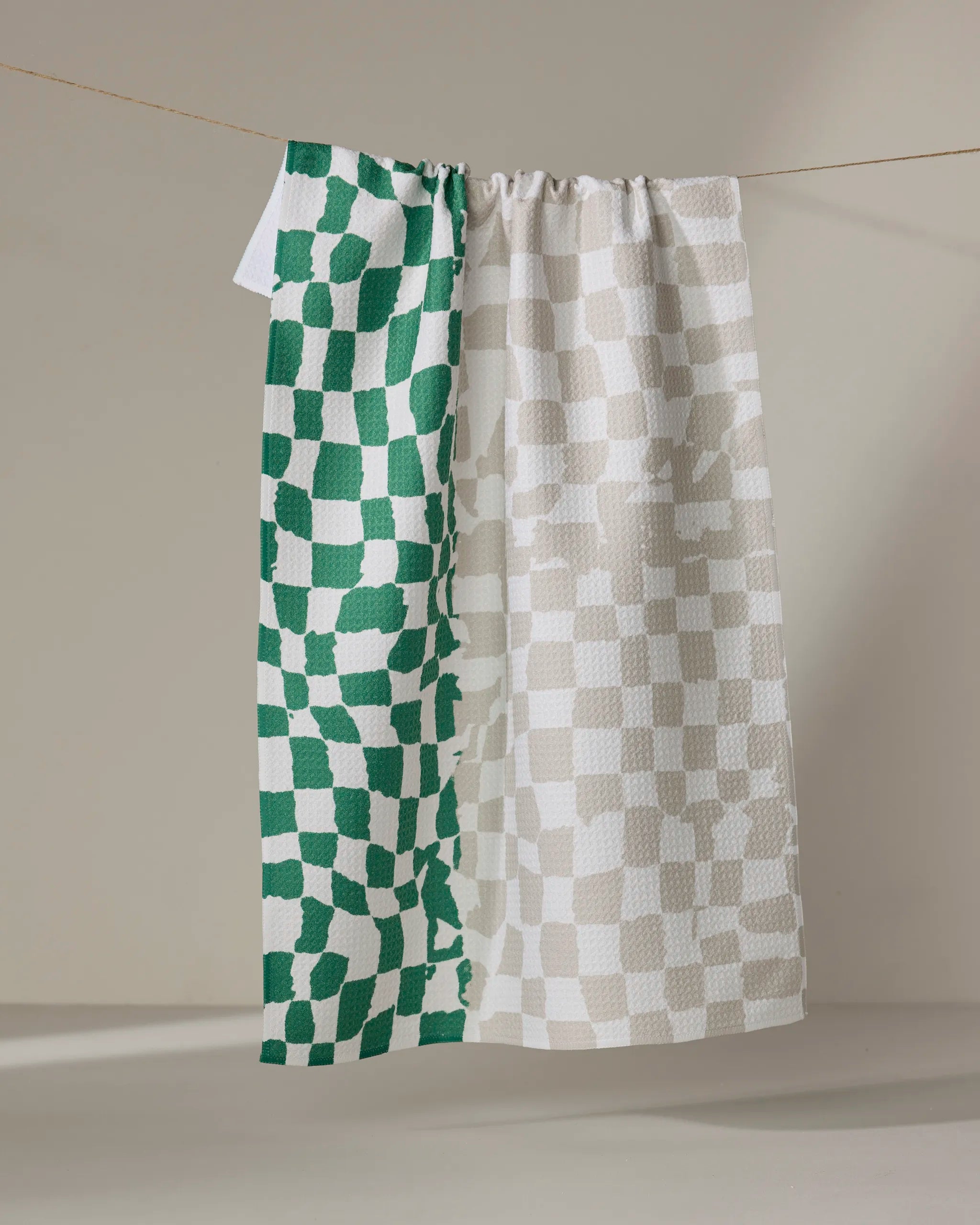 The Dual tea towel by Geometry, made from recycled materials, hangs on a neutral background. Its left side displays green and white uneven checks, while the right side features beige and white checks in a checkered pattern.