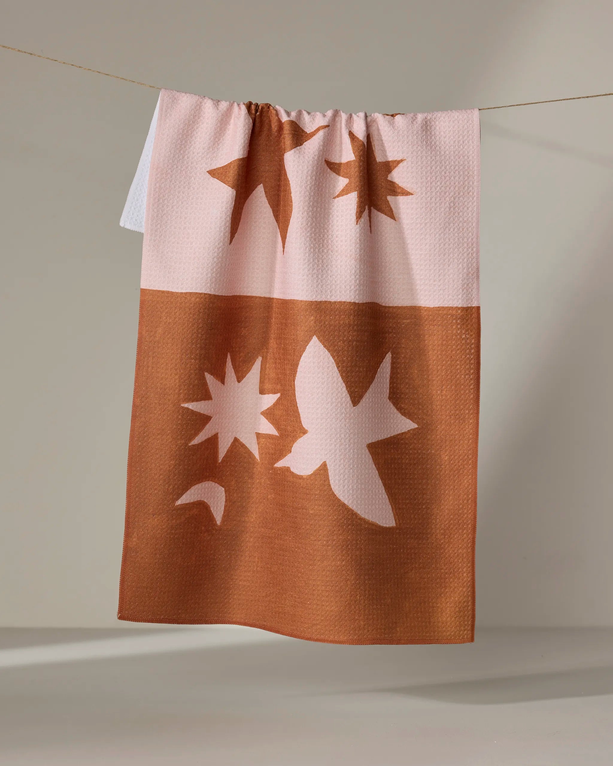 Jen B. Peters' "Duality" tea towel, made from recycled materials, is quick-drying and two-toned in pink and brown. It displays abstract designs—stars and a bird—in contrasting colors on each half, perfect for stylish drying.