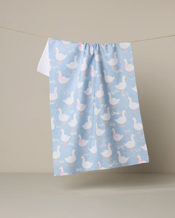 The Geometry "Duck Trail" tea towel, in light blue with a quick-drying fabric and a repeating pattern of white ducks—some with pink bows—hangs on a line against a beige background.