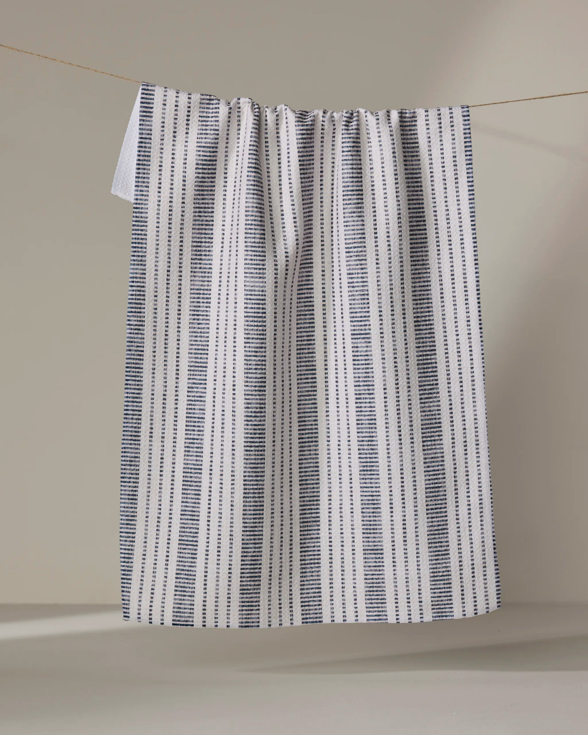 The "Dyer Blue" tea towel by Becki Owens, featuring vertical navy stripes on white cotton, hangs gathered at the top against a light background, showcasing its ultra-absorbent design.