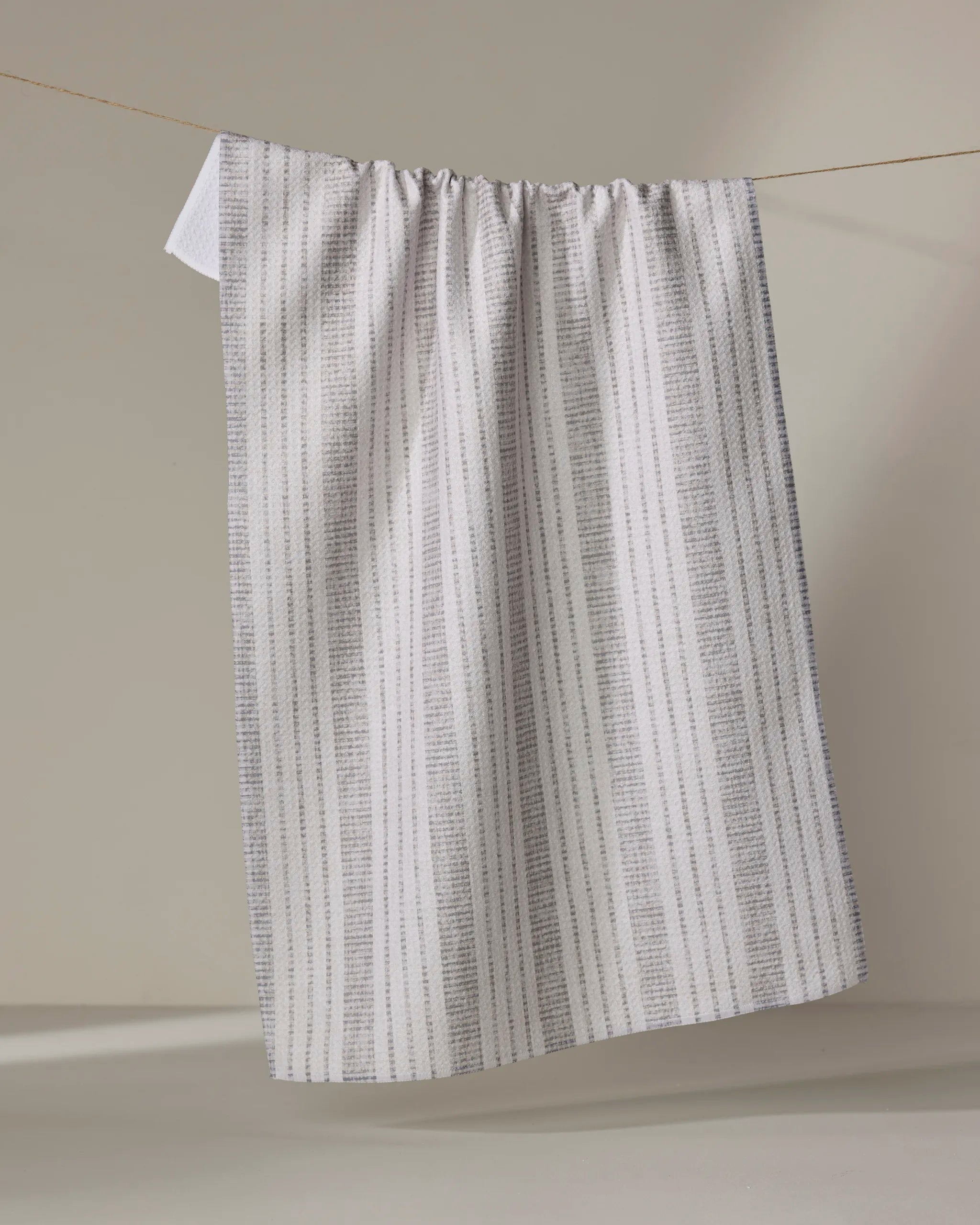 The Becki Owens "Dyer Grey" towel, featuring thin gray vertical stripes on white, hangs on a clothesline against a neutral beige background.