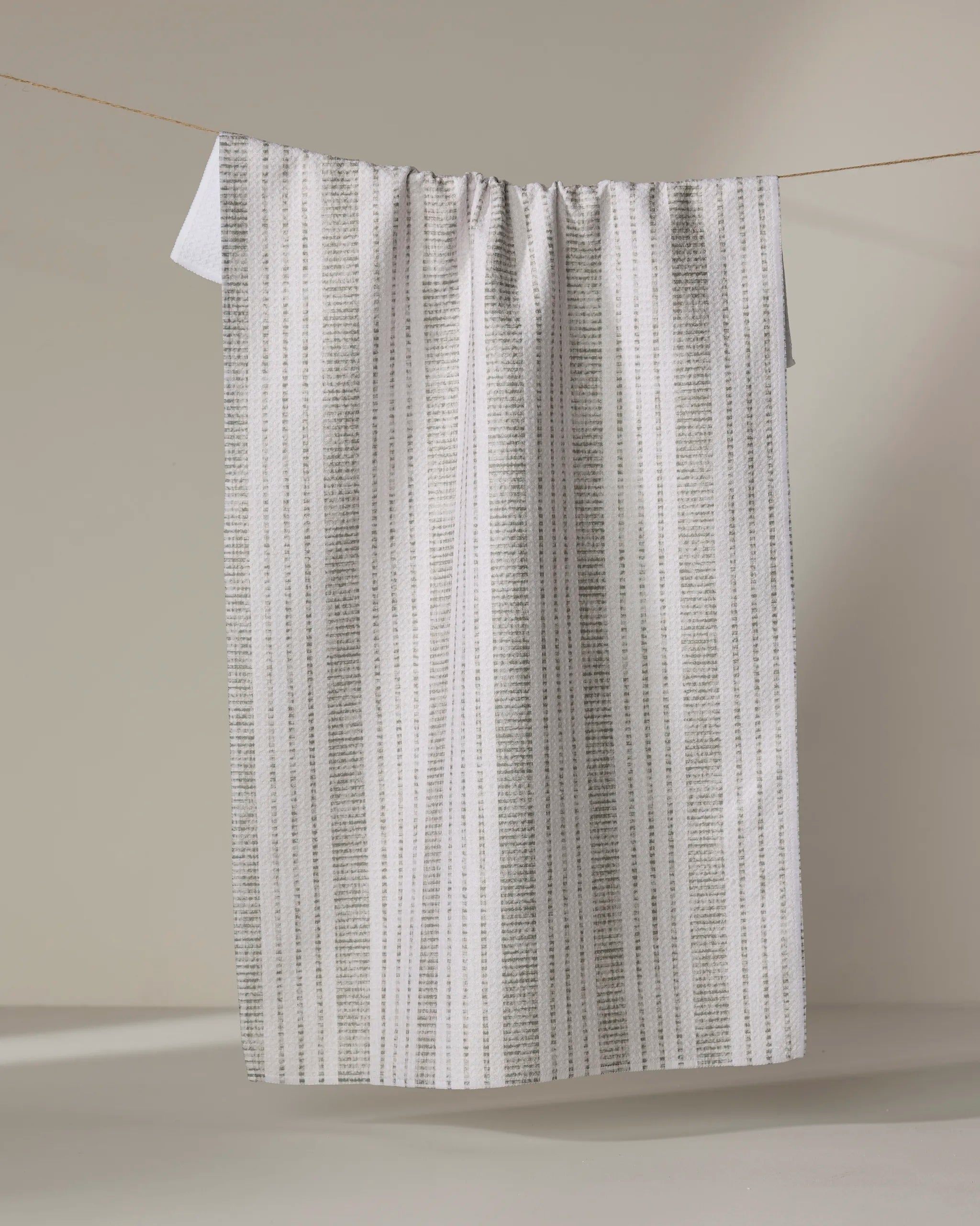 The Becki Owens Dyer Sage tea towel, featuring thin vertical gray stripes on white and made from recycled materials, hangs on a clothesline against a plain beige background.