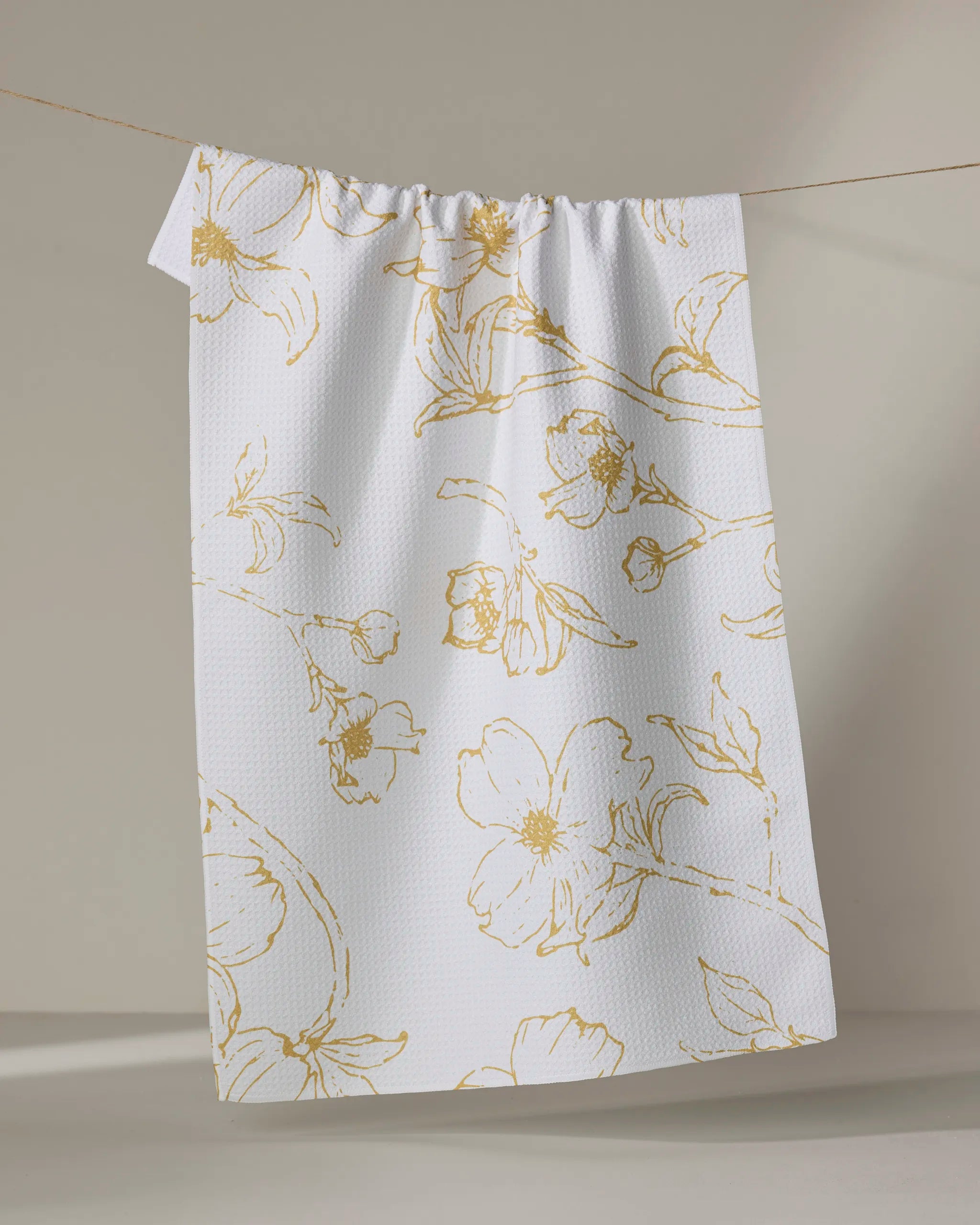 The Early Blossoms tea towel by Ashley Celena features a white textured fabric with a gold floral pattern, combining style and absorbency for versatile kitchen use.