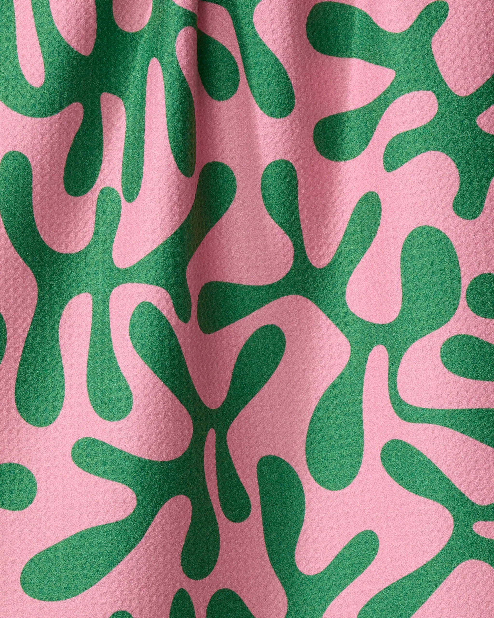 A close-up of the Early Morning Plants tea towel by Marina Solodka shows textured, ultra-absorbent fabric with green abstract shapes on pink. Soft folds and shadows highlight its recycled material and organic design.
