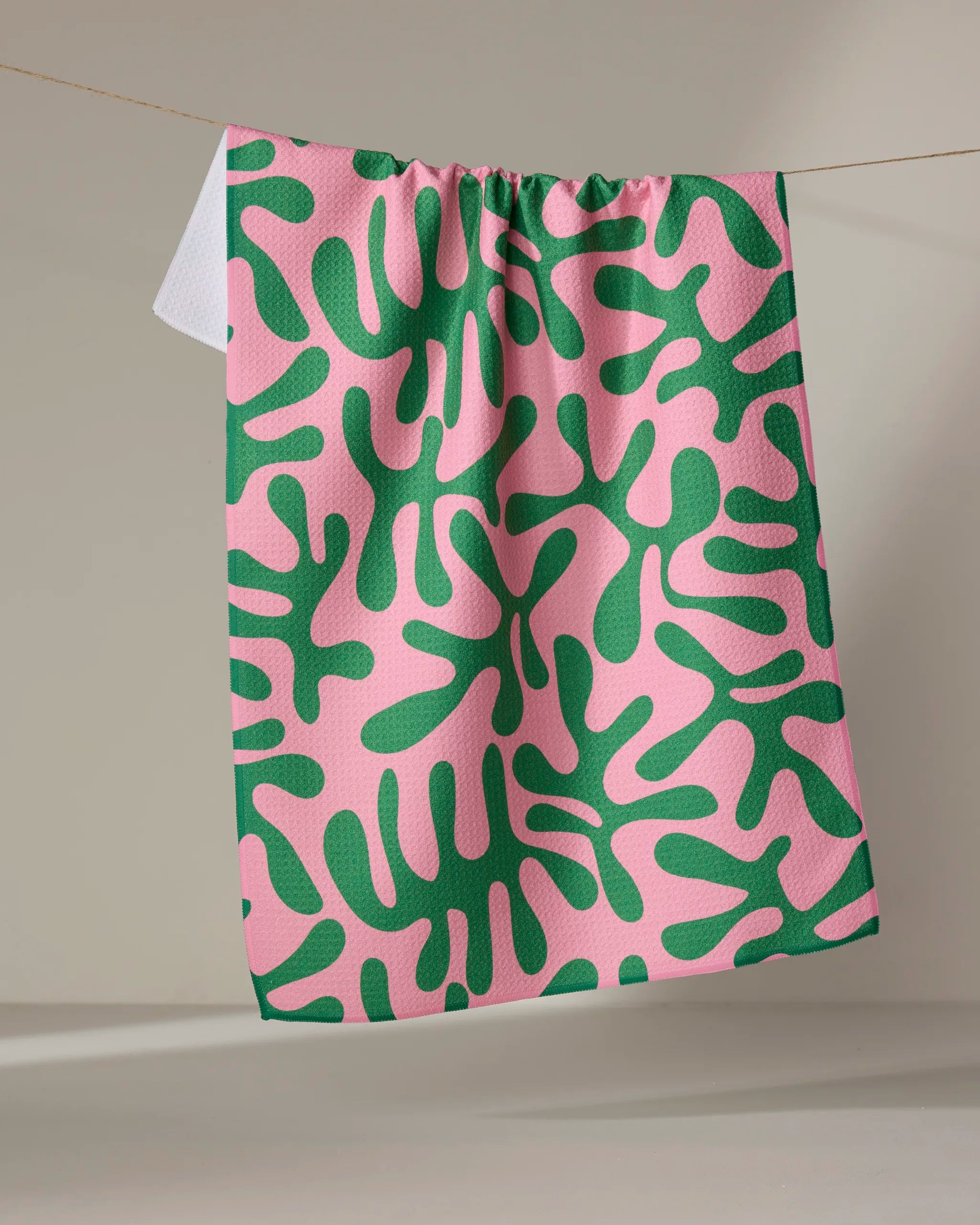 The "Early Morning Plants" tea towel by Marina Solodka, featuring a bold green abstract pattern on pink, hangs from a clothesline against a neutral backdrop.