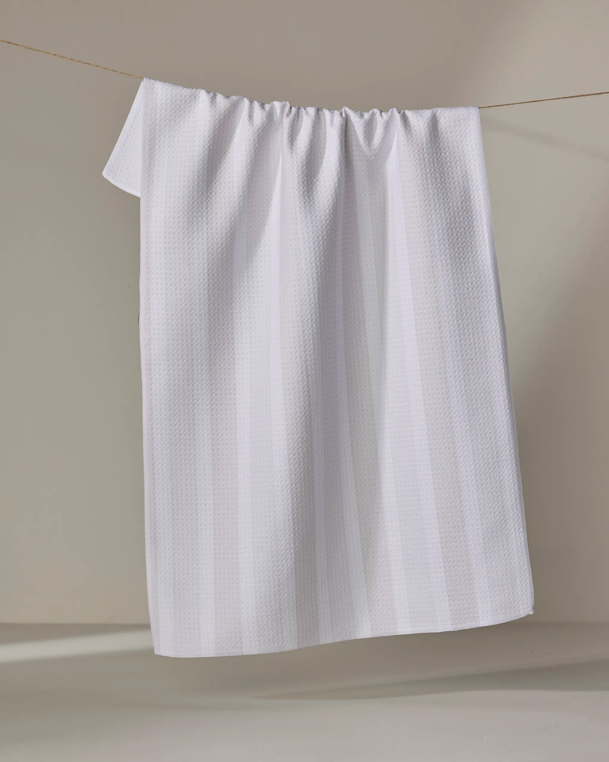 The Buy Guide’s Earn Your Stripes - Beige textured kitchen towel hangs neatly on a clothesline against a neutral backdrop, with soft lighting enhancing its woven pattern and subtle shadows.