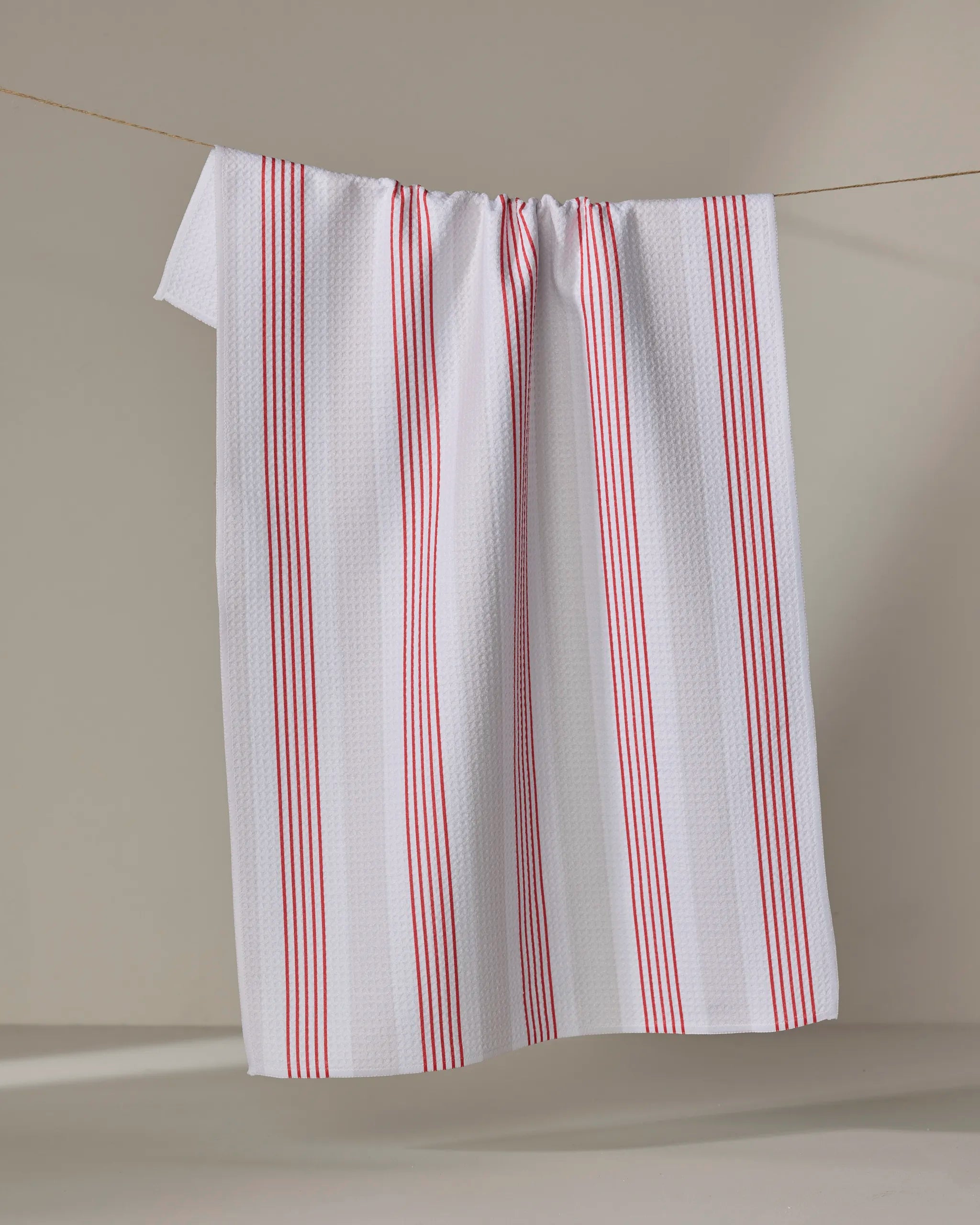 The Buy Guide’s "Earn Your Stripes - Candy Apple" is a white, recycled tea towel with red vertical stripes, shown hanging neatly on a thin clothesline against a plain, light-colored background.