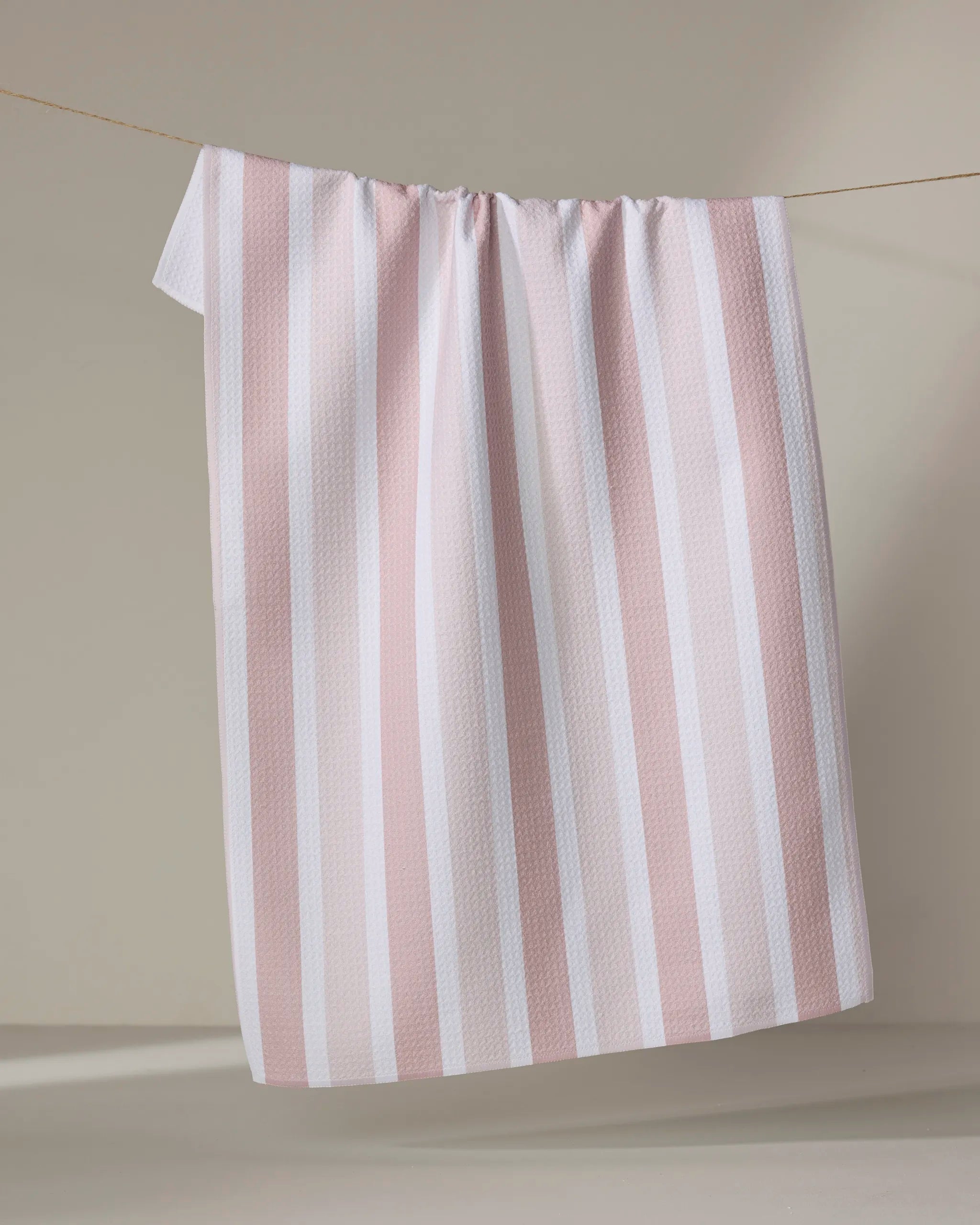 The Buy Guide's Earn Your Stripes - Dusty Rose tea towel, with dusty rose and white vertical stripes and a textured, waffle-like fabric, hangs draped over a thin line against a neutral beige background.