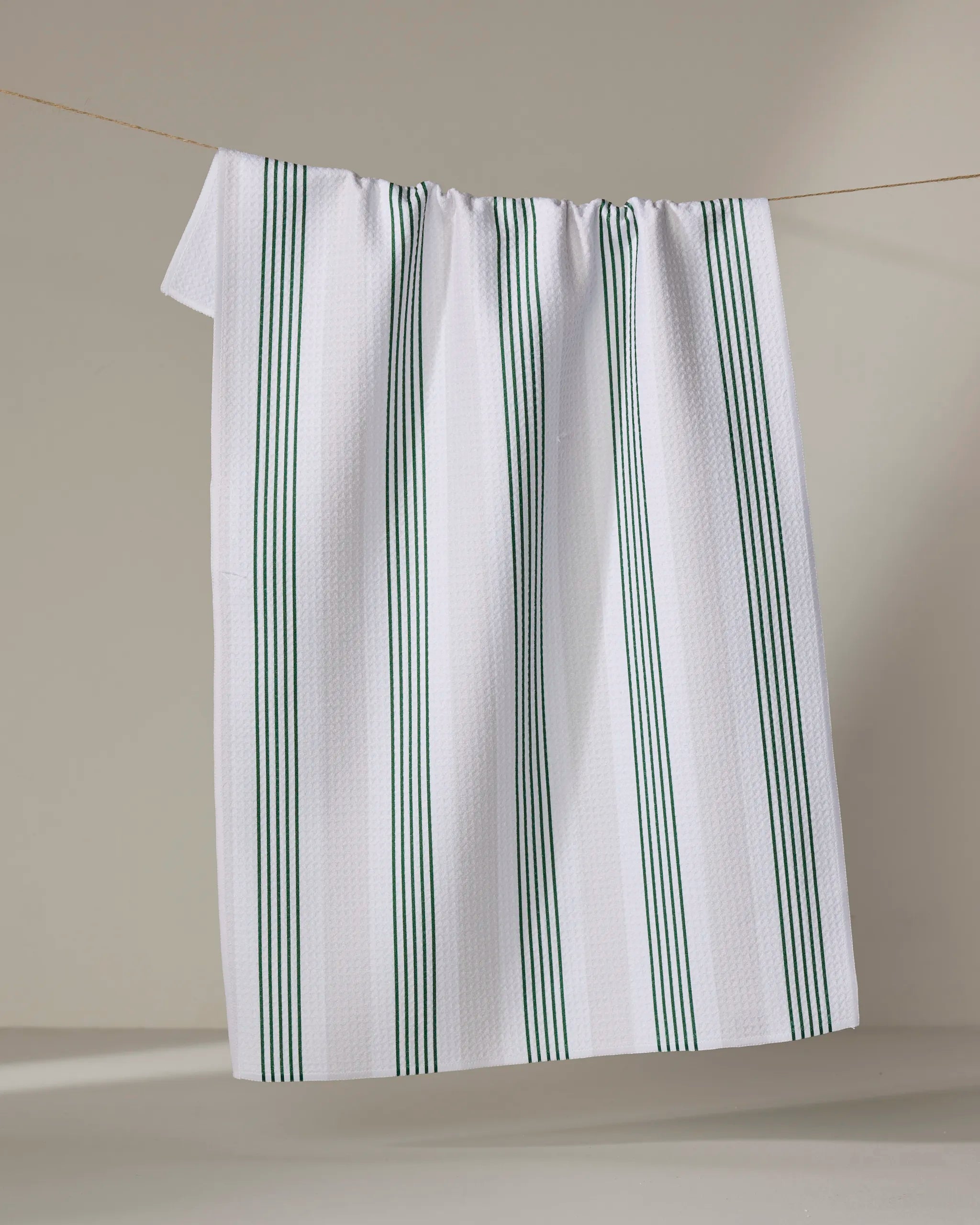 The Buy Guide's "Earn Your Stripes - Evergreen" white tea towel with vertical green stripes, made from recycled materials, hangs on a clothesline against a plain, light background.