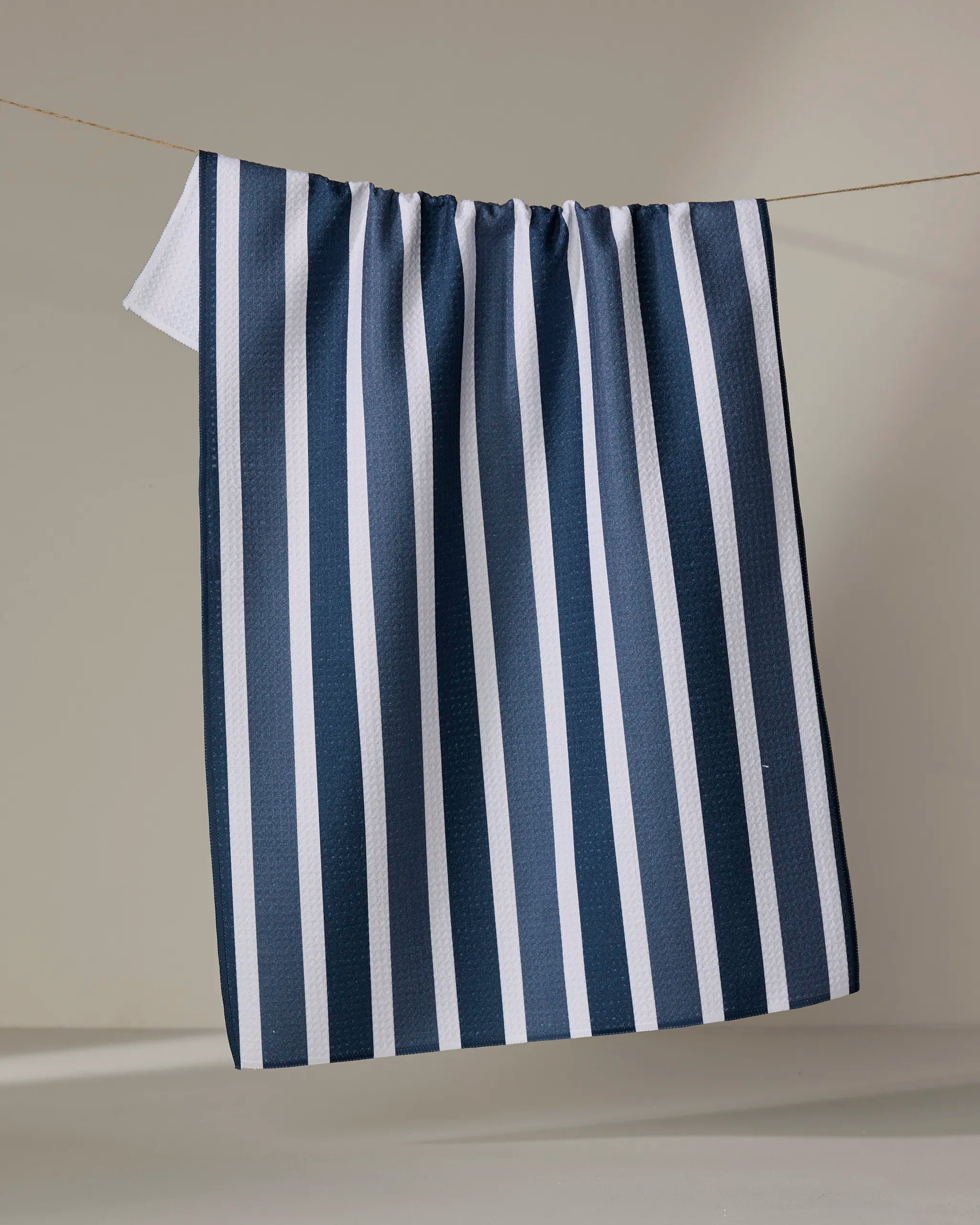 The Buy Guide’s Earn Your Stripes - Navy tea towel features blue and white vertical stripes, hanging on a thin, light-colored string against a neutral background.