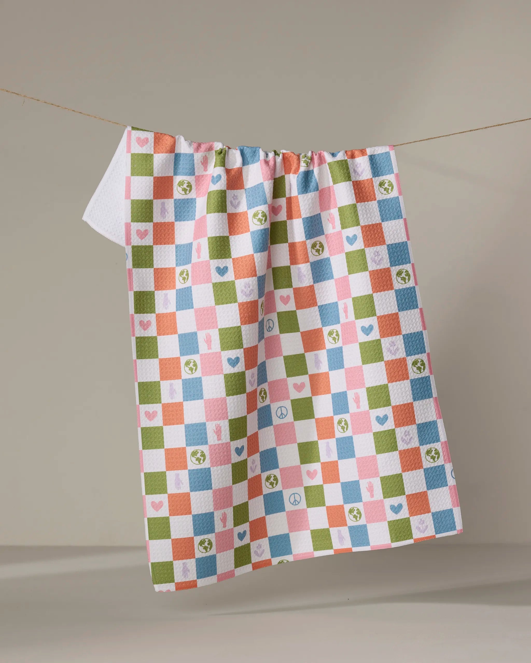 The Kayla Ann Earth Day Checkers tea towel hangs on a line, showcasing pink, blue, green, and orange squares with hearts, peace signs, and Earth icons. Made from recycled materials.