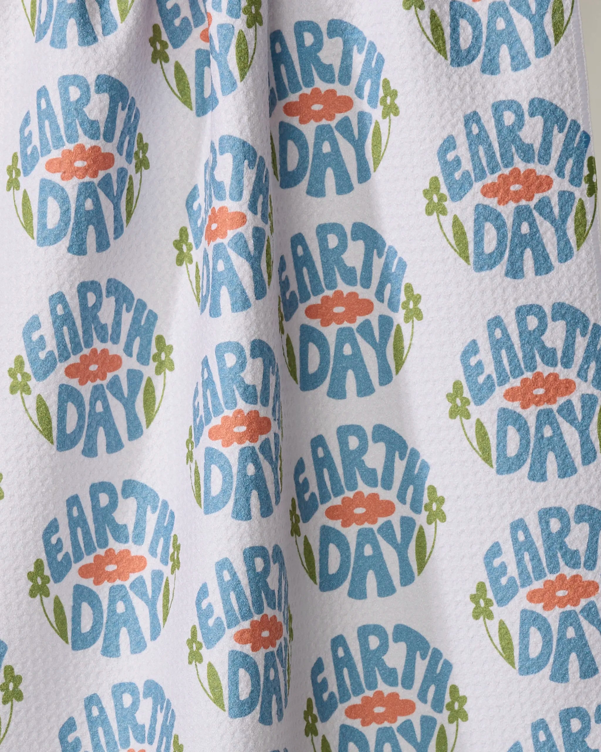 The Earth Day Stamp tea towel by Kayla Ann features blue "EARTH DAY" text surrounded by orange flowers with green leaves and stems, all on textured white ultra-absorbent recycled material.